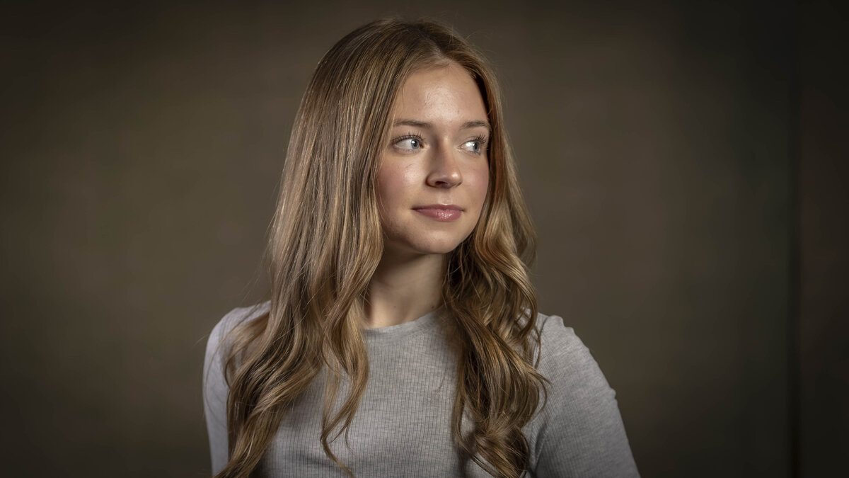 Professional headshot in CT of a young woman with long brown hair wearing a fitted white top, softly lit against a dark background, by Rob Faber Photography, specializing in headshots in CT and headshots in MA.
