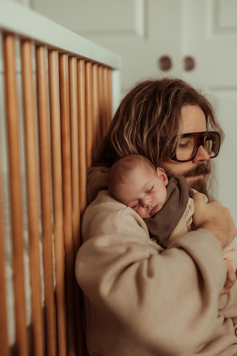 Close up of Dad sitting against baby's crib while holding his new son over his shoulder