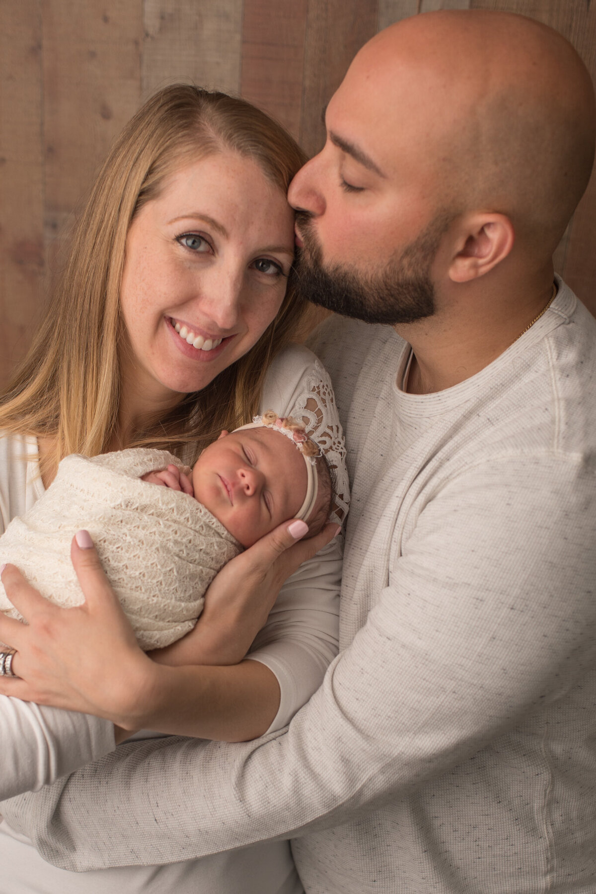 Mother and father in white, holding newborn daughter wrapped in white, father kissing mother's head