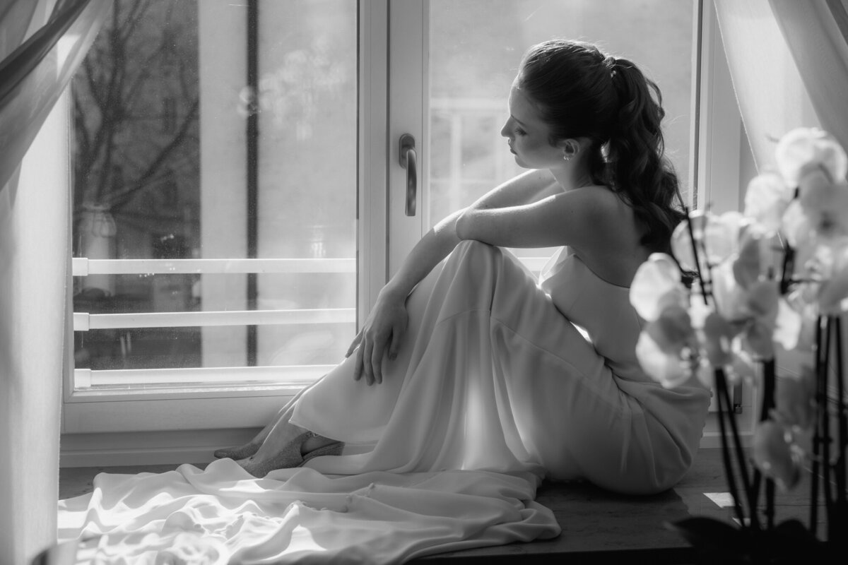 Das Brautmodel sitzt mit angewinkelten Knien auf einem Fensterbrett und blickt hinab.