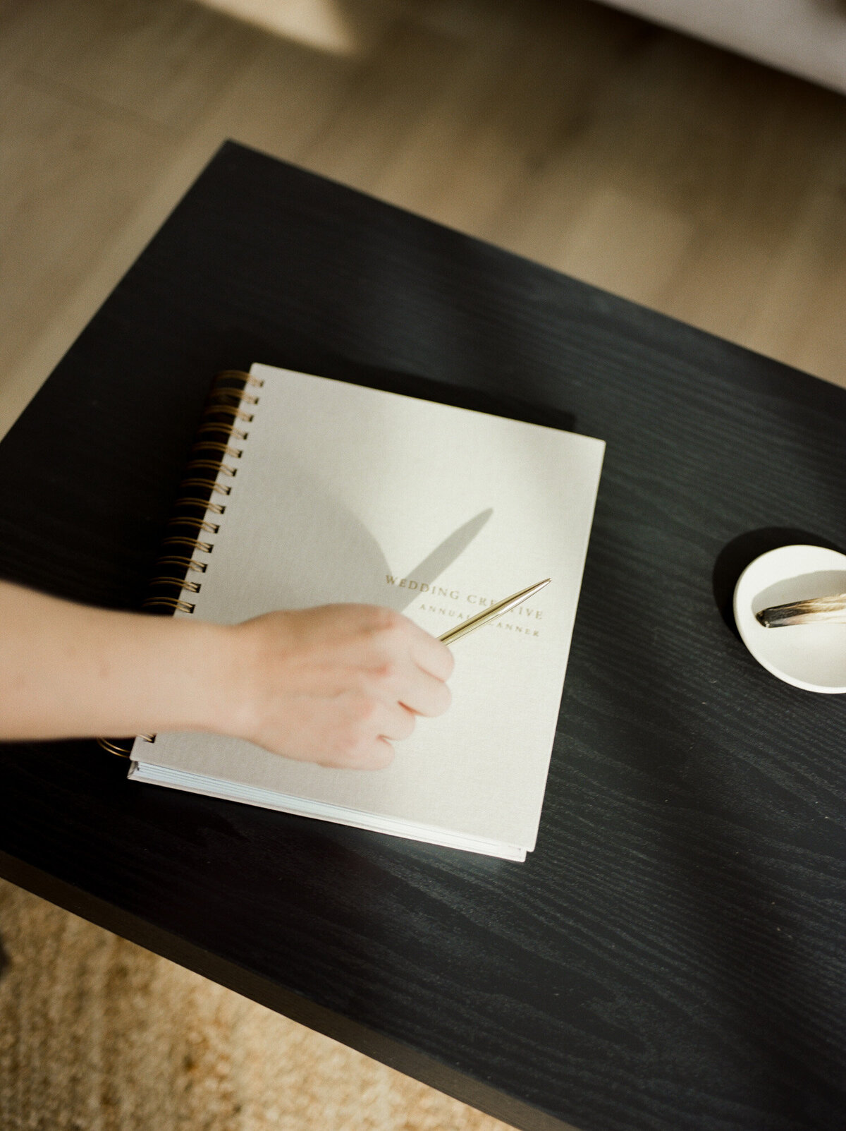 hand reaches for gold pen, casting shadow over beige linen planner with gold lettering on black table