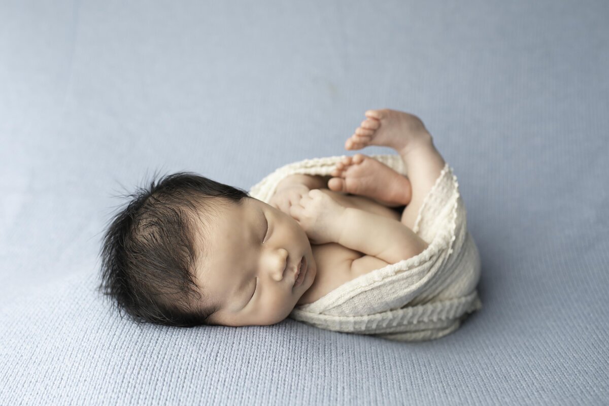 Newborn baby boy with a full head of dark hair, snugly wrapped in a white swaddle, peacefully resting on a soft blue backdrop