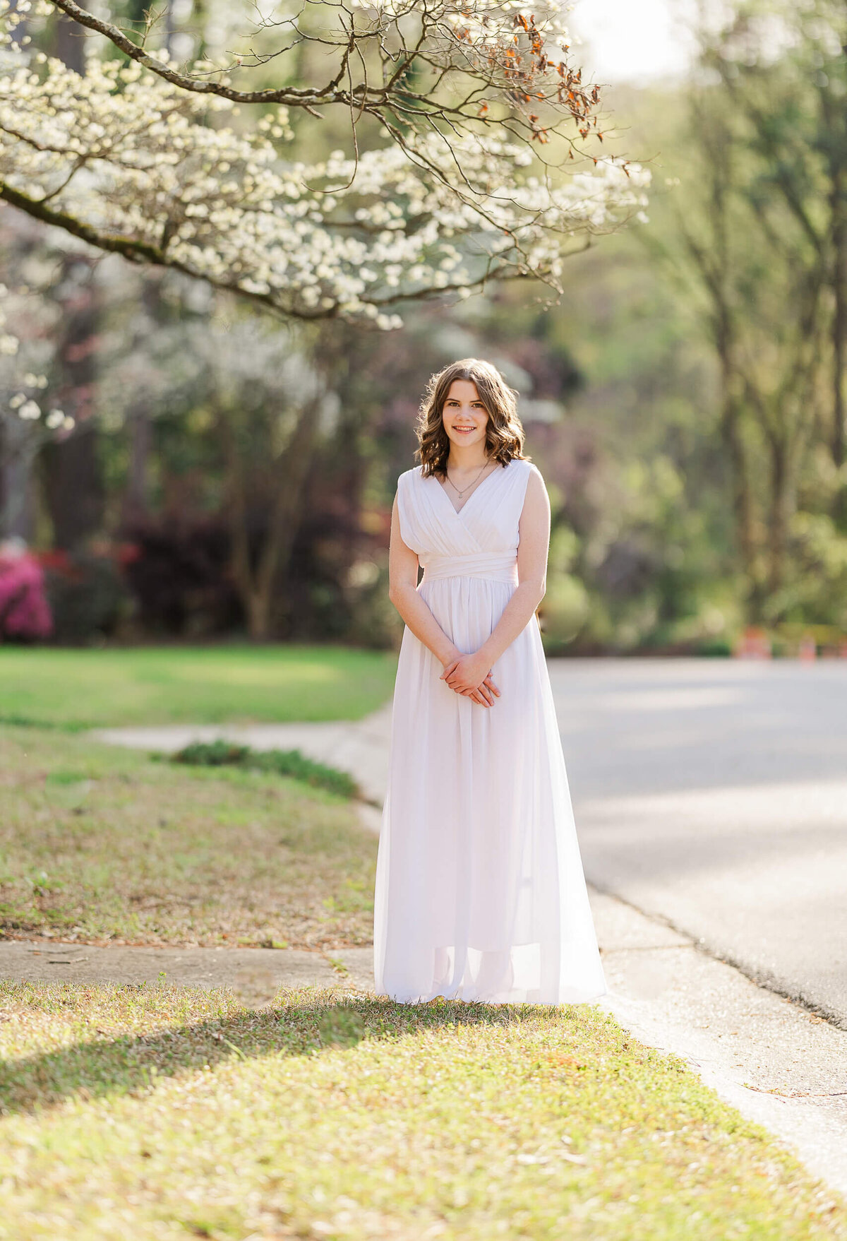 A woman in a long white dress stands on a sidewalk in a sunlit outdoor setting. Trees with blossoms and greenery surround her, reminiscent of a Fayetteville NC family photographer's perfect setting, as the road curves in the background. The scene evokes a sense of spring or early summer.