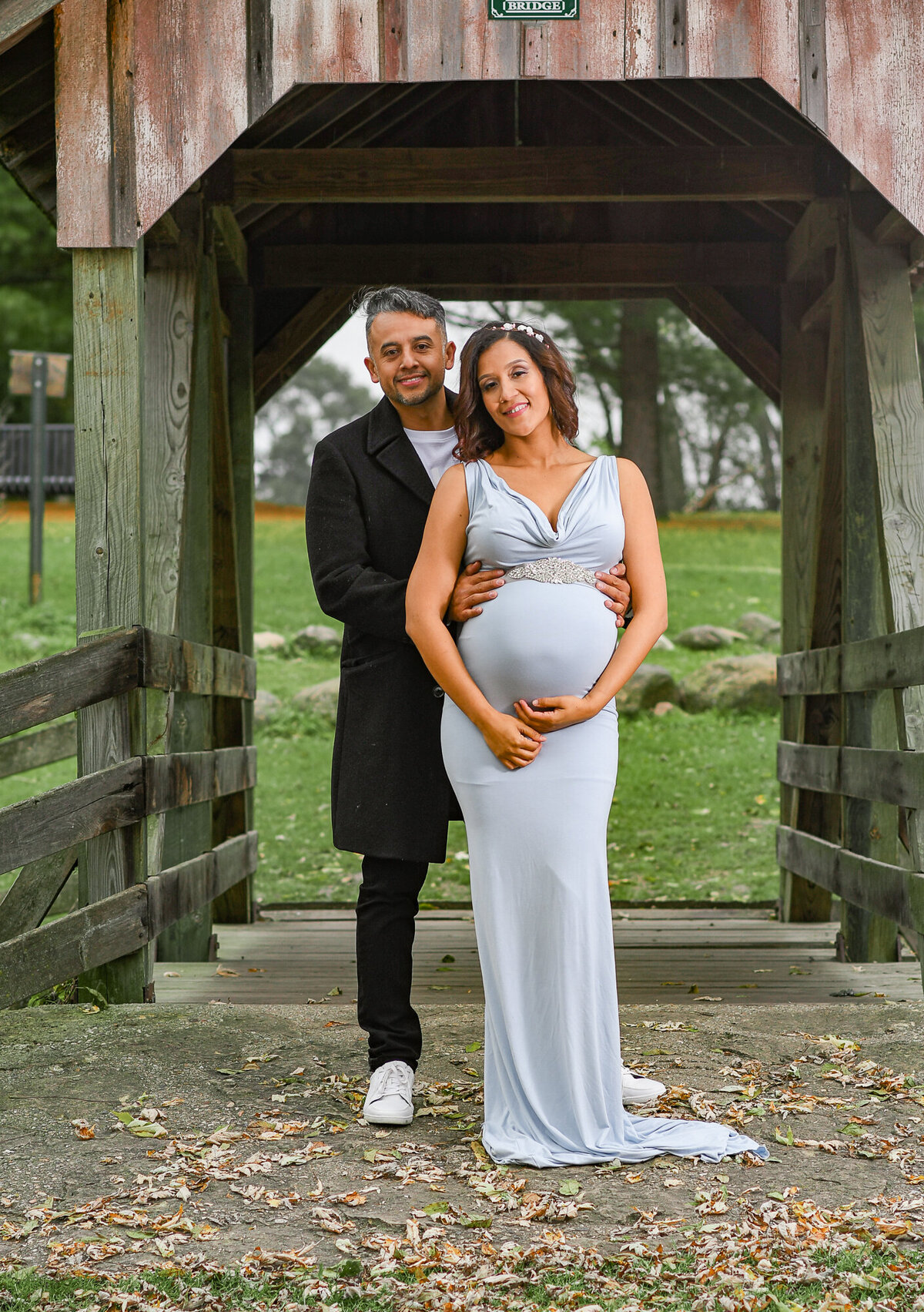 Man wearing a long black coat standing on a covered bridge with his pregnant wife smiling at the camera