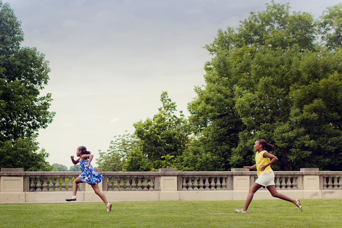Two girls running in the grass at the upper level of Newfields in Indiana. One has on a bright blue dress, the other as a yellow top.