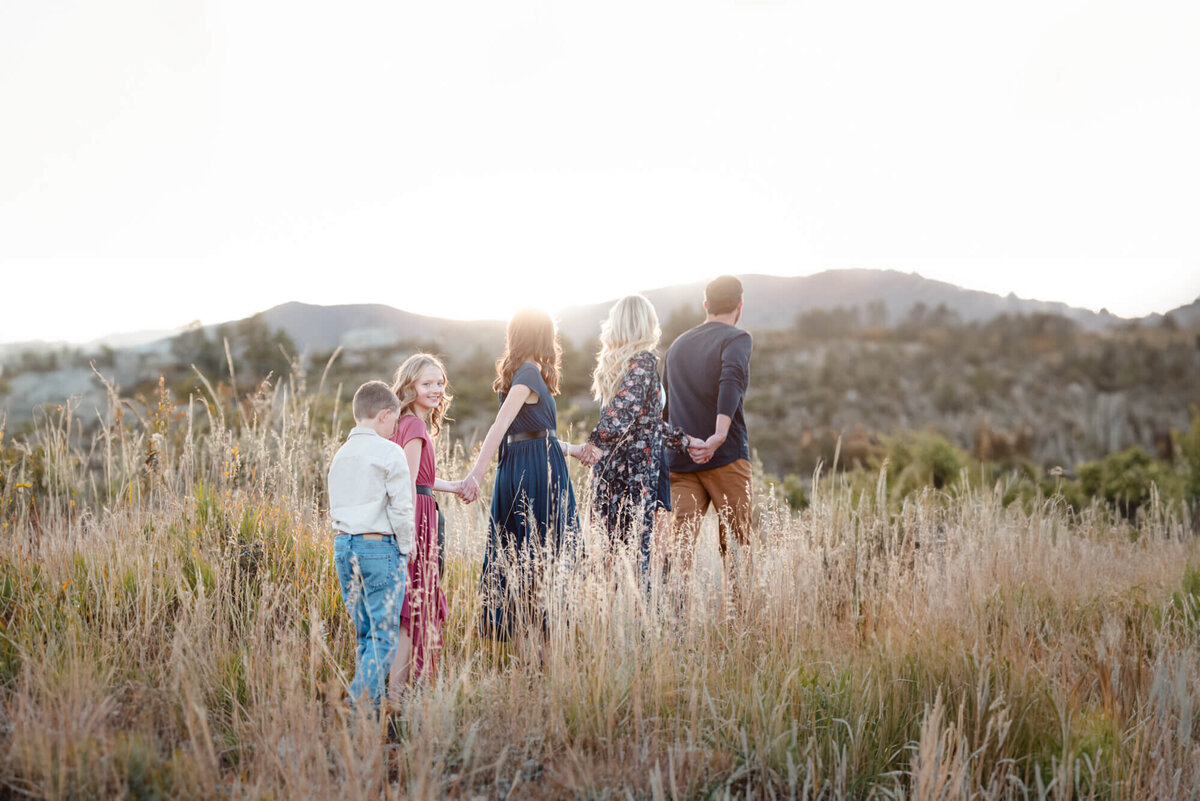 A father leads his family of 5 through a grassy trail at sunset while they all hold hands for a Denver family photographer