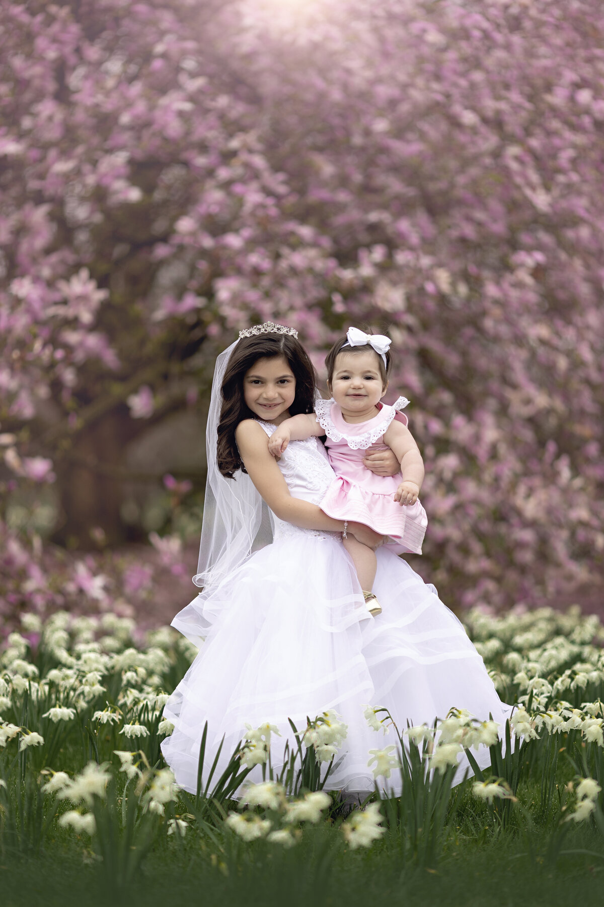 A young girl in a white dress and tiara holds her toddler baby sister in a pink dress in a field of white wildflowers