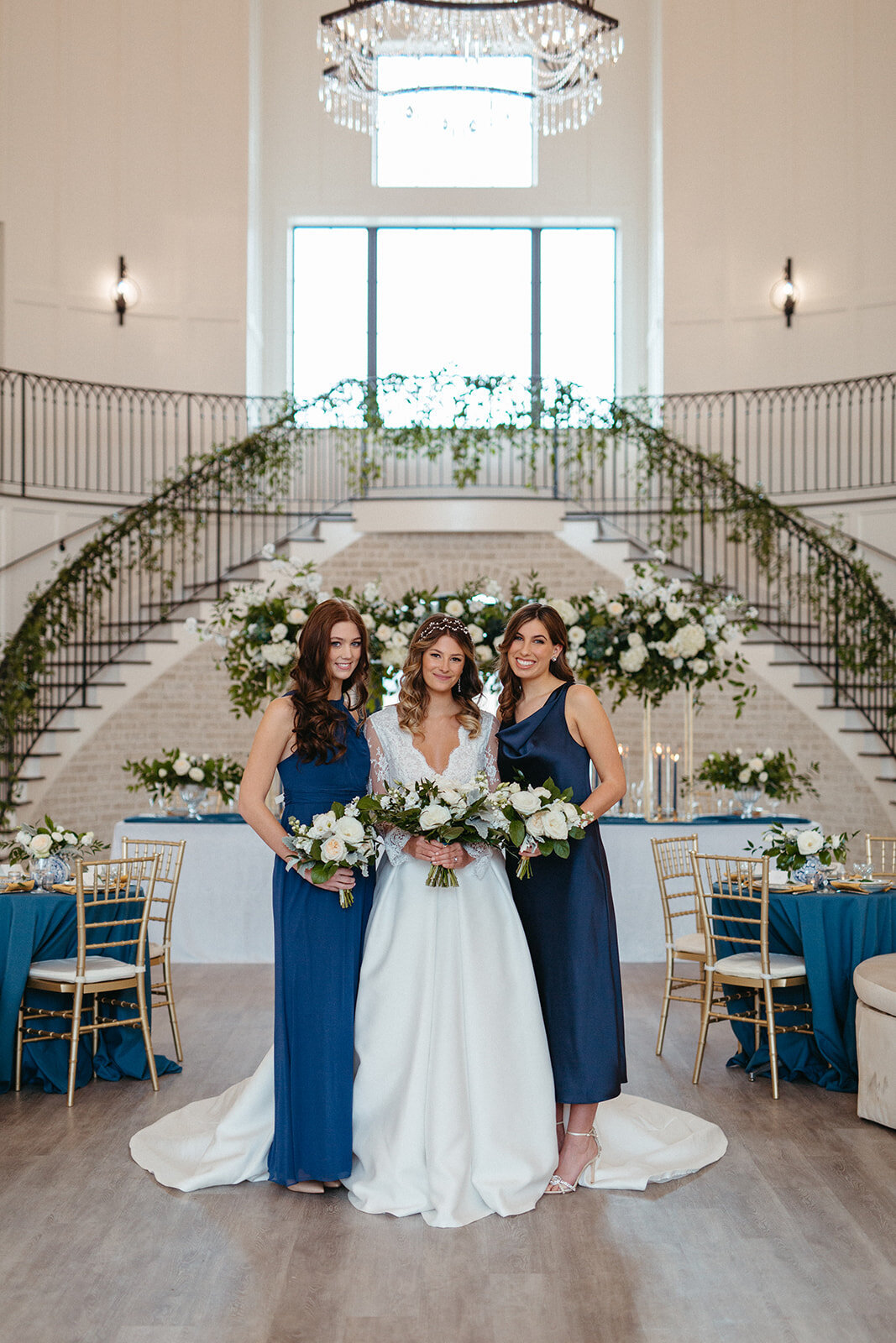 A bride in a white wedding gown and two bridesmaids in blue gowns holding white bouquets in a banquet room.