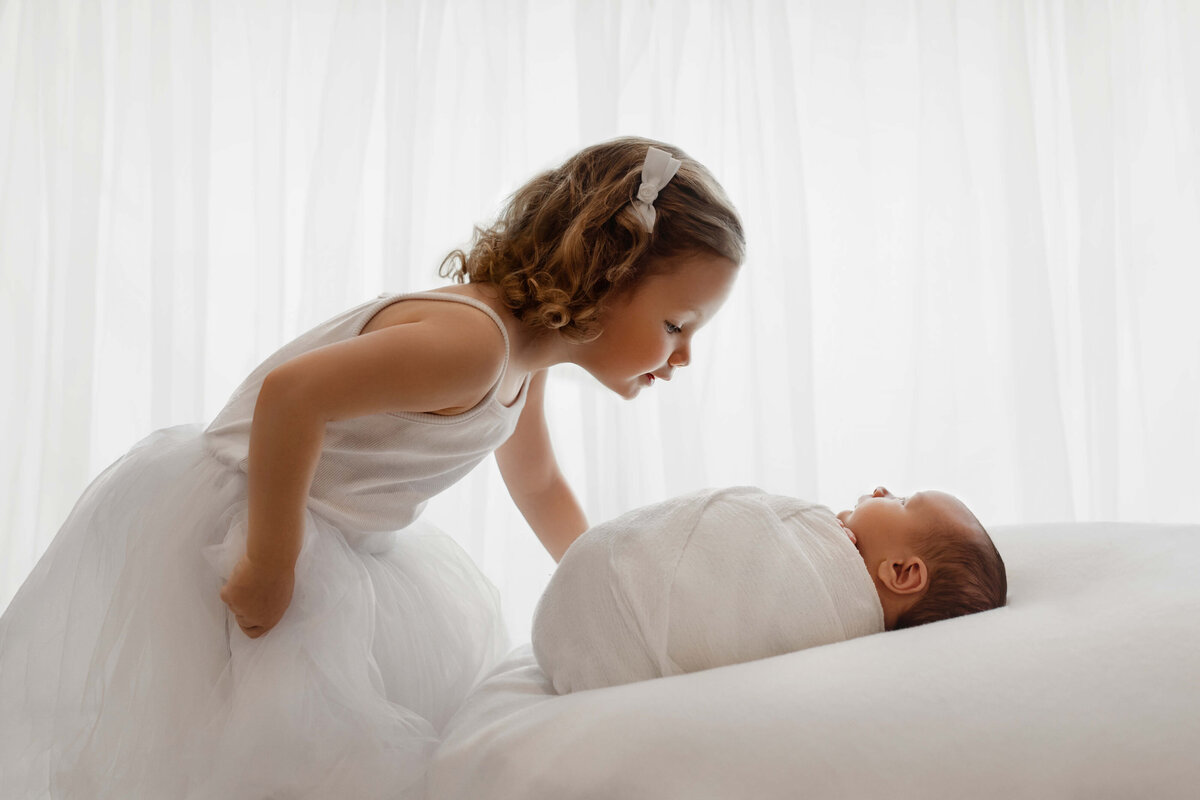big sister bending down to kiss her newborn brother  wearing a white dress in front of back-lit white curtains