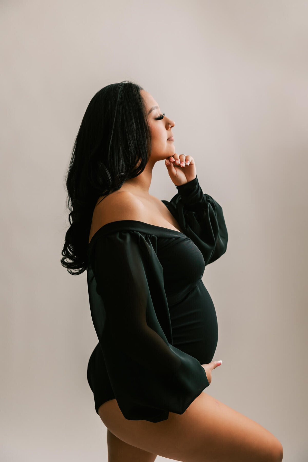 portrait of a pregnant woman wearing black bodysuit. She has one hand under belly and the other under her chin and long black hair.
