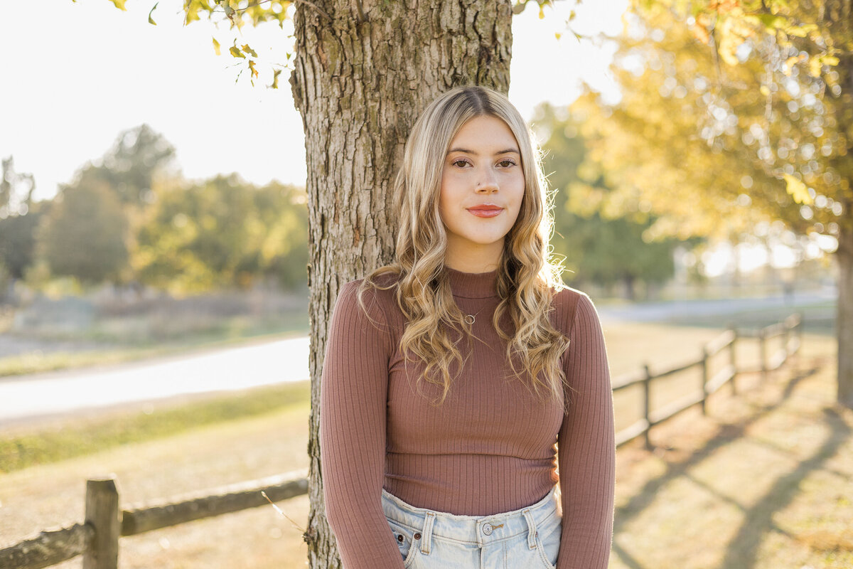High school senior female with long blonde hair, standing against a tree with the sun and trees behind her, wearing a long sleeve purple shirt, blue jeans, and looking at the camera