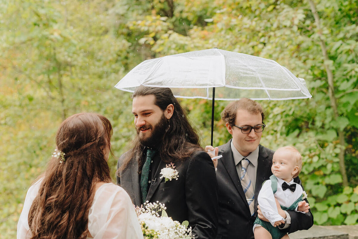 Bride and groom looking at each other while guest holds baby and umbrella