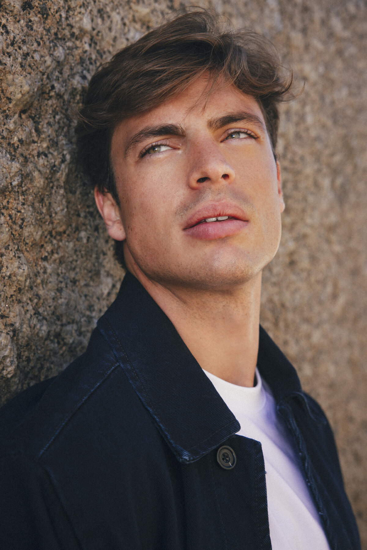 Close-up portrait of male model with tousled brown hair leaning against a textured rock wall in Cape Town. Wearing a dark jacket and white T-shirt, he gazes slightly upward with soft sunlight highlighting his face.