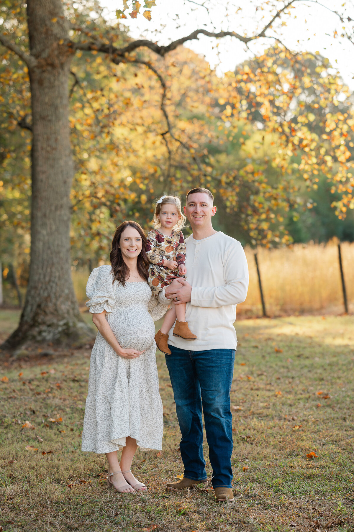 Pregnant mother holds her belly while her husband stands beside her holding their 3 year old little girl. They smile at the camera while the sun sets at Prospect Farms in Lawrenceville. Maternity photos by Amanda Touchstone