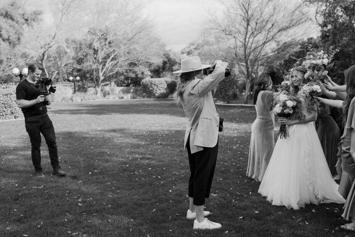 Husband and wife photo and video team capturing a wedding day. A black and white photo, the husband and wife photographer and videographer are shooting a bride and her bridesmaids before the wedding ceremony.