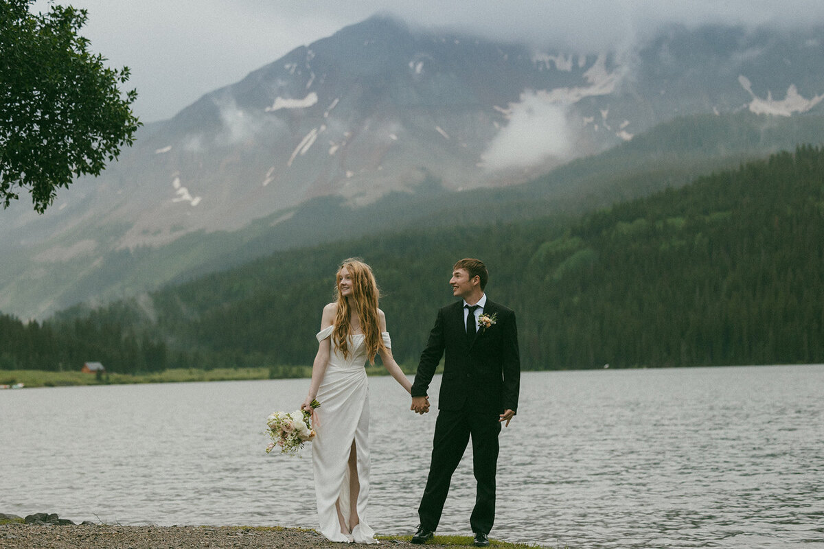 bride and groom holding hands in front of lake and mountains cloudy sky