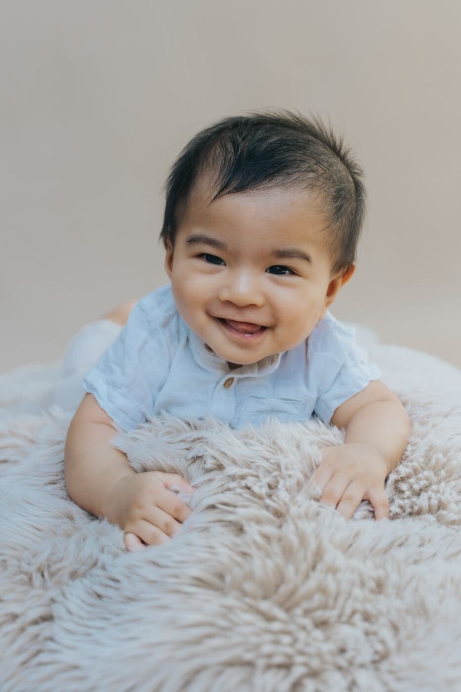 Smiling baby boy lying on a fluffy beige blanket, wearing a light blue shirt and looking directly at the camera.