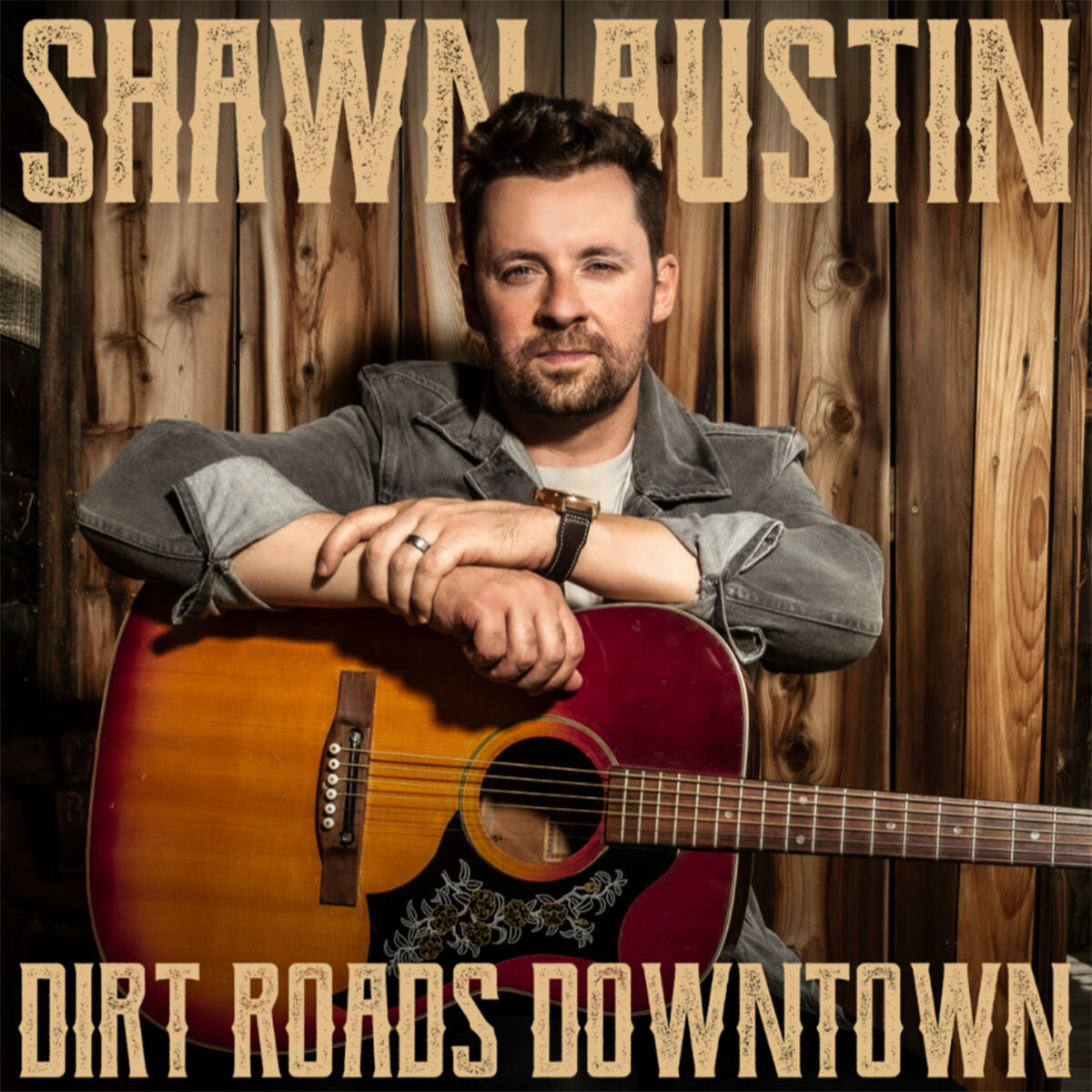 Country artist Shawn Austin single cover Dirt Roads Downtown sitting with guitar in front of wood wall