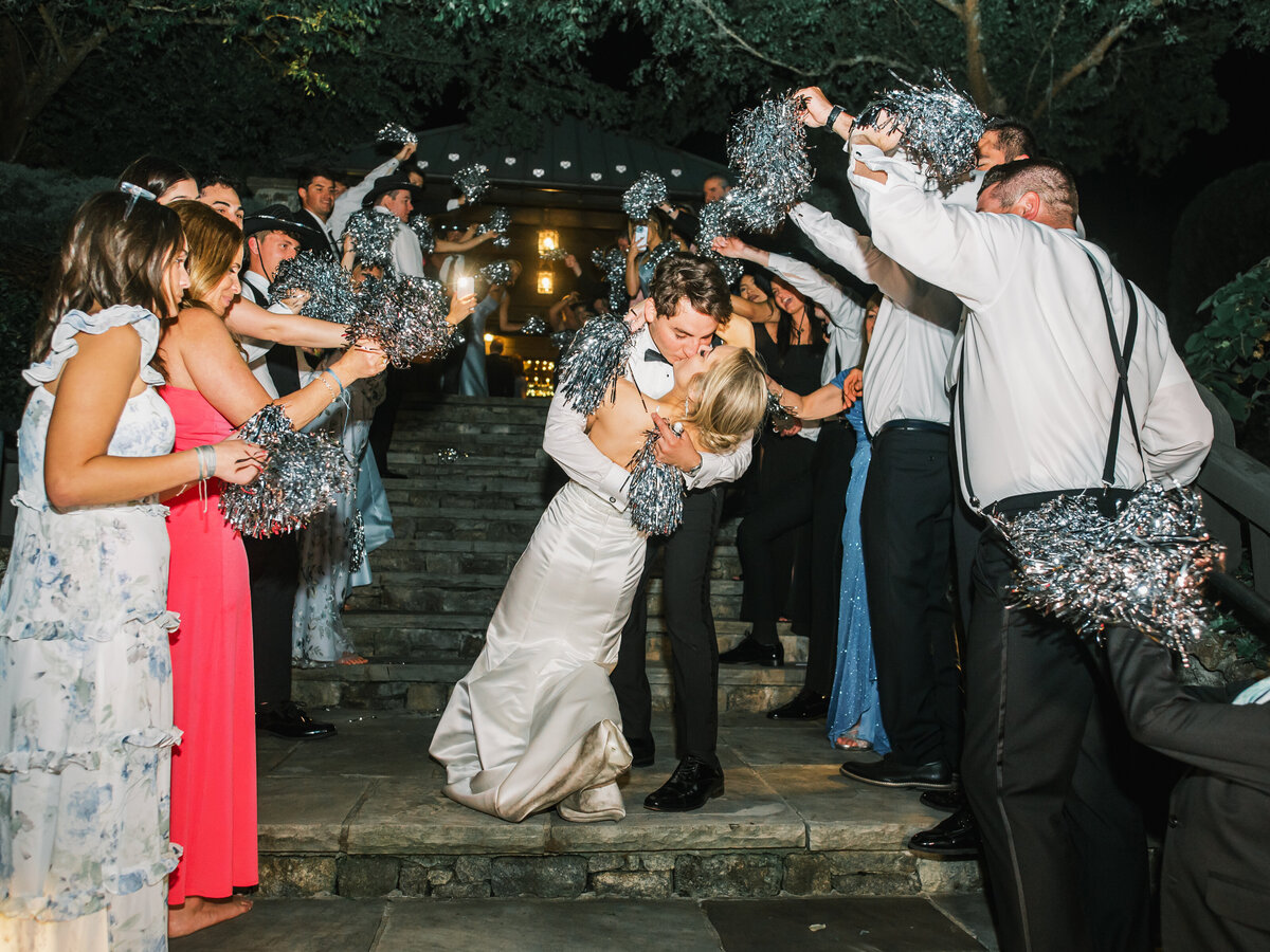 Bride and groom share a kiss during their grand exit with guests cheering and waving silver pom-poms at Old Edwards Inn in Highlands, North Carolina.