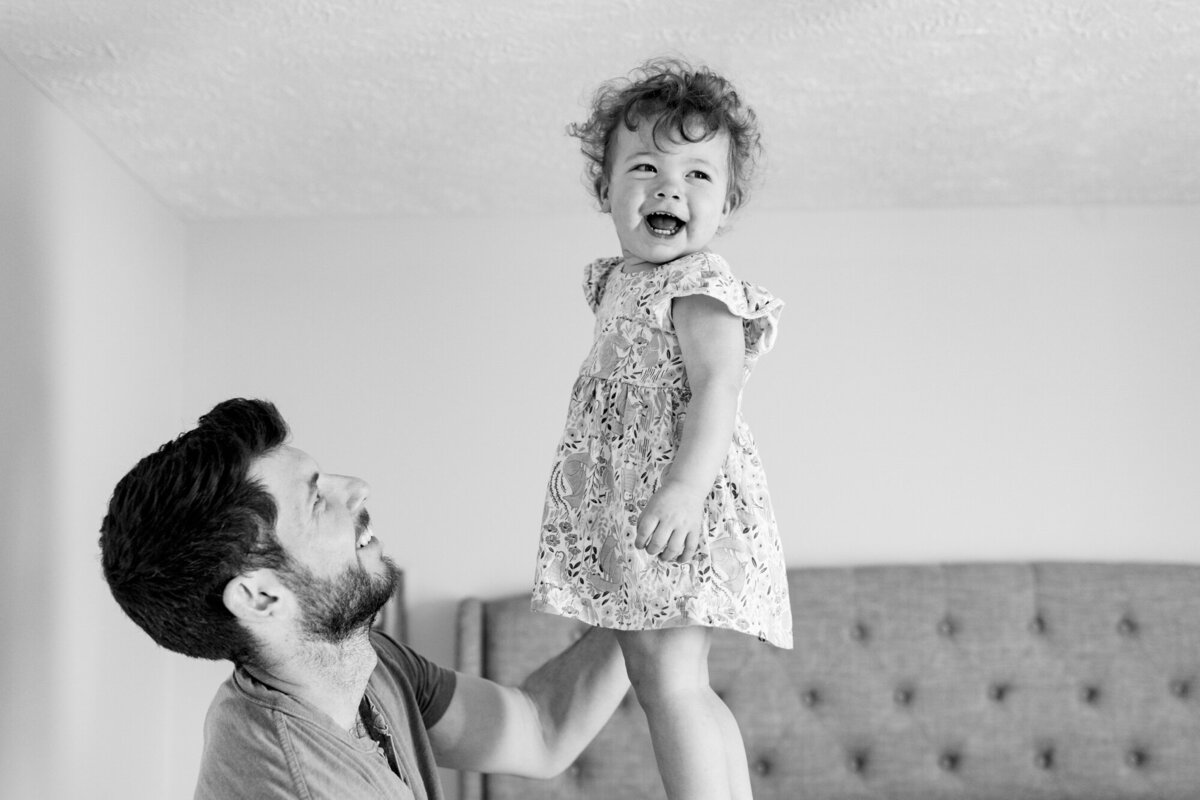 a dad balances his little girl in one hand above the bed in case she falls. The little girl looks over at her mom while giggling.