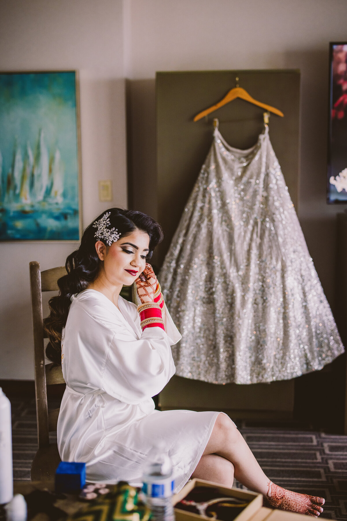 A sikh bride in a robe puts on earrings before her wedding reception with her silver lehnga hanging in the background