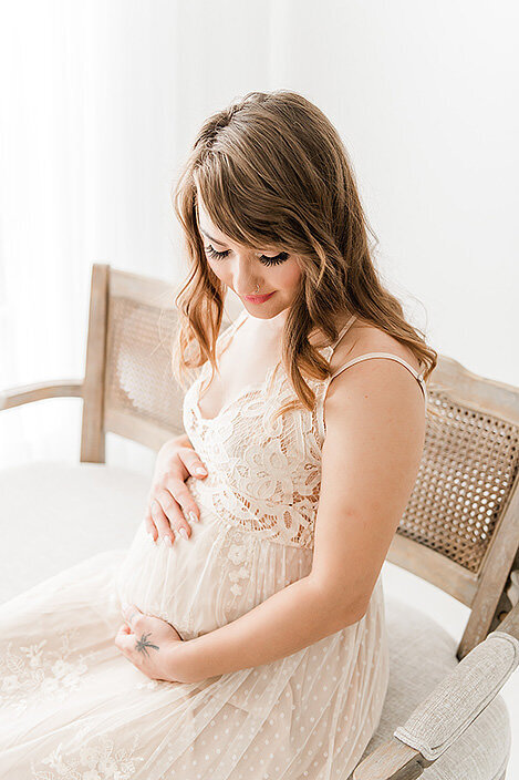 Glowing mom-to-be in a blush lace gown sits quietly, gazing at her belly with a soft smile in a bright white room.