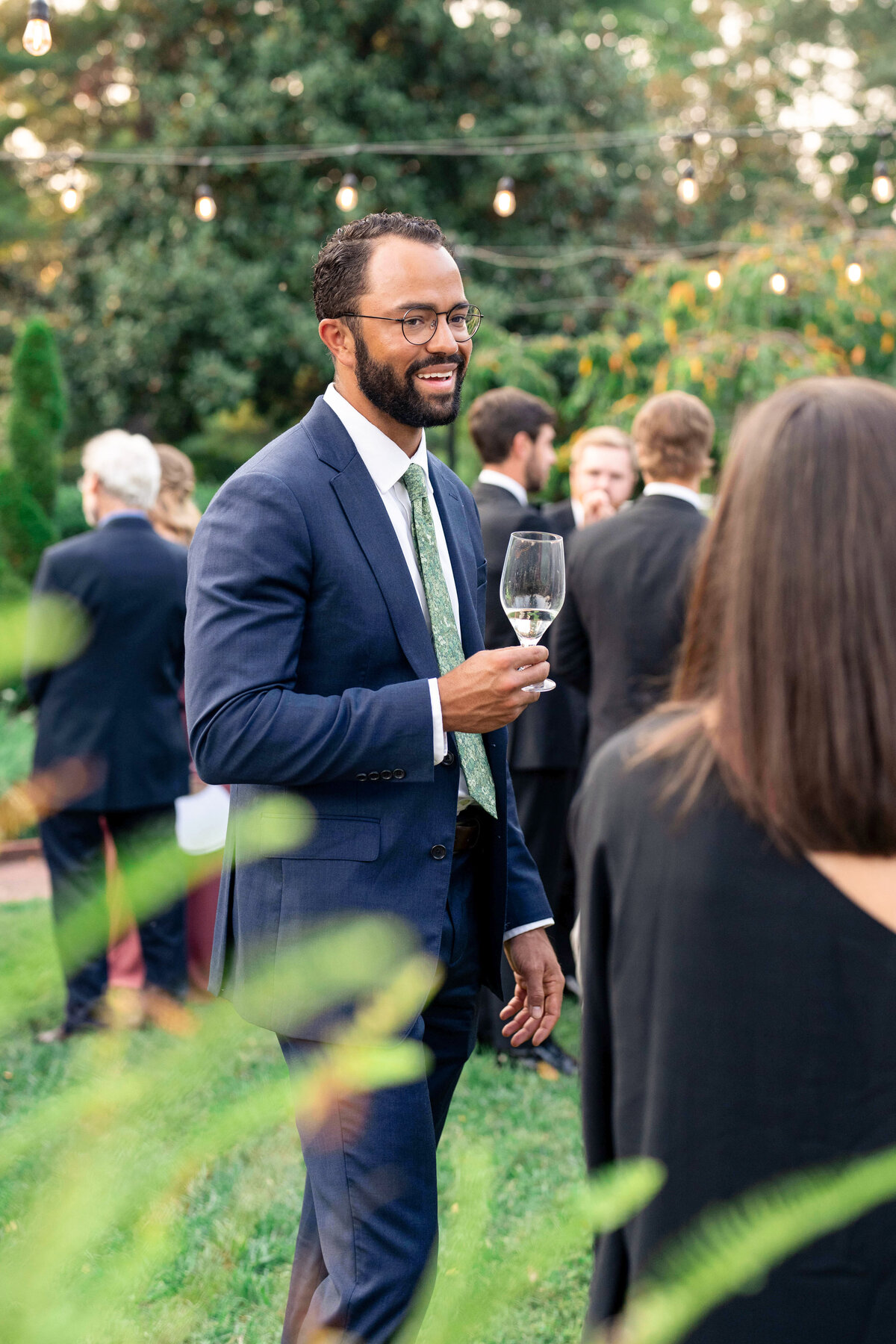A man in a blue suit with a green tie holds a wine glass while smiling at an outdoor social event. Other attendees are in the background, and the setting includes greenery and string lights.
