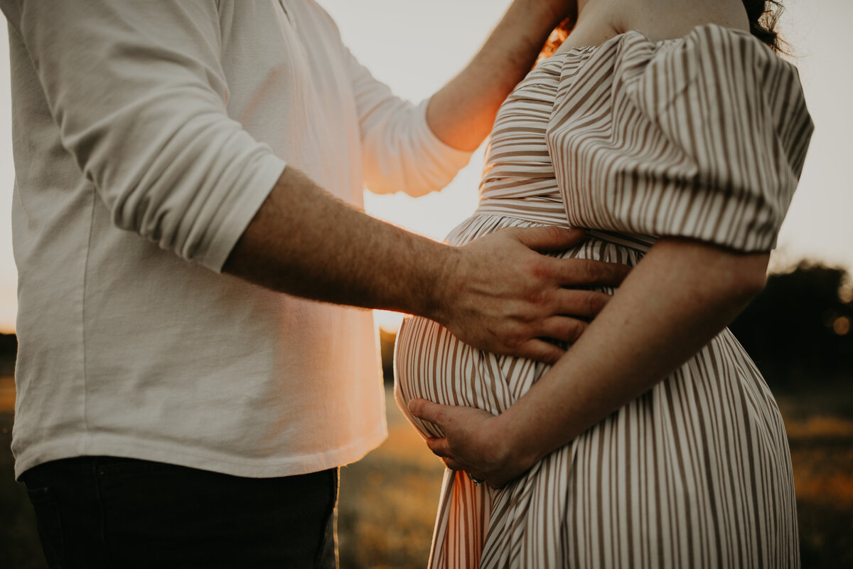 Expecting couple hold mother's pregnant belly while the sun peeps through during maternity session with maternity photographer Cameron Fox of Foxc Photography in Blum, Texas.