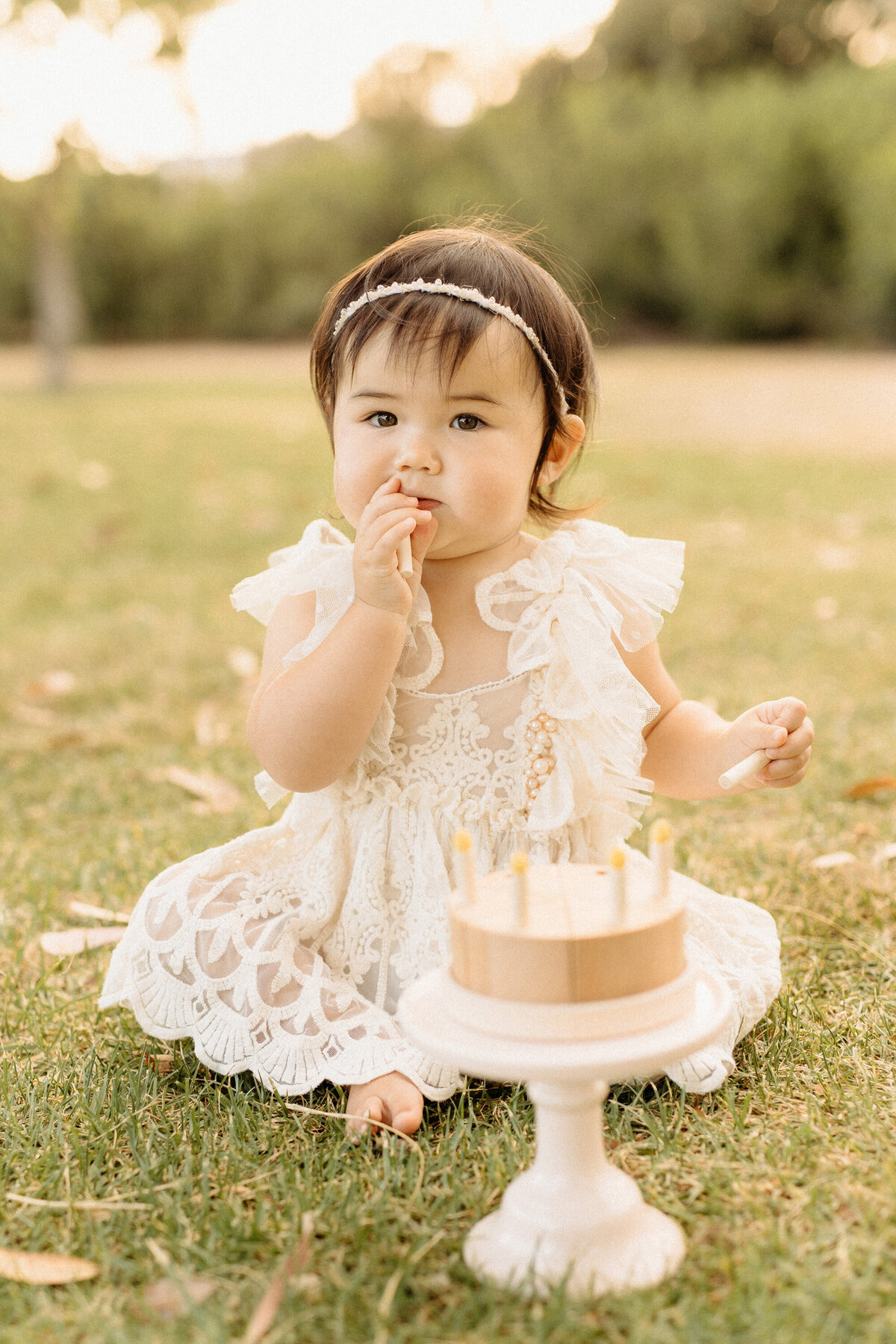 Adorable 1 year old baby girl in a lace dress enjoying her first birthday cake smash outdoors, photographed in natural light by San Diego baby photographer Yuka Schad.