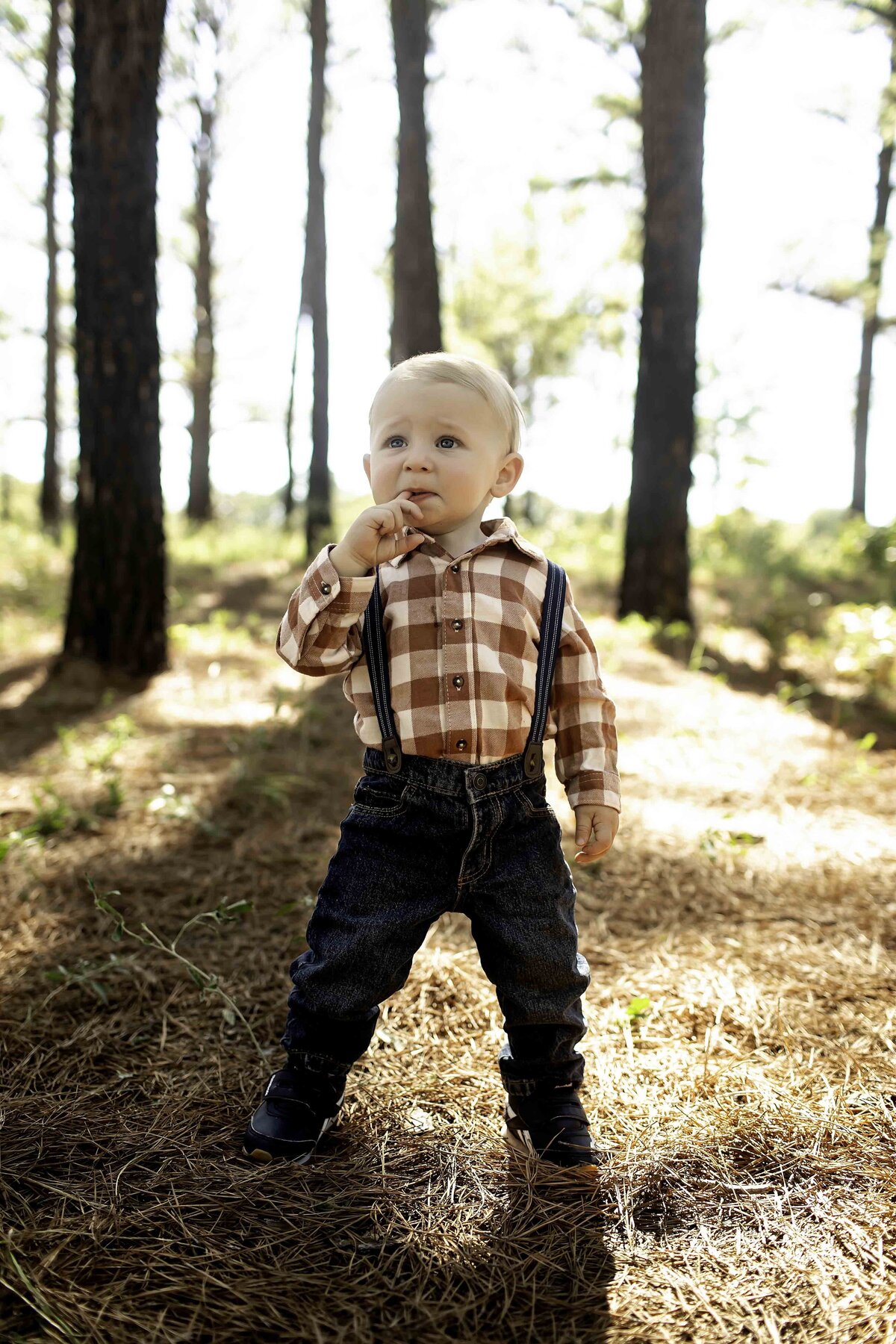 Birthday boy in suspenders and overalls, playfully posing among pine trees, with the sunlight filtering through