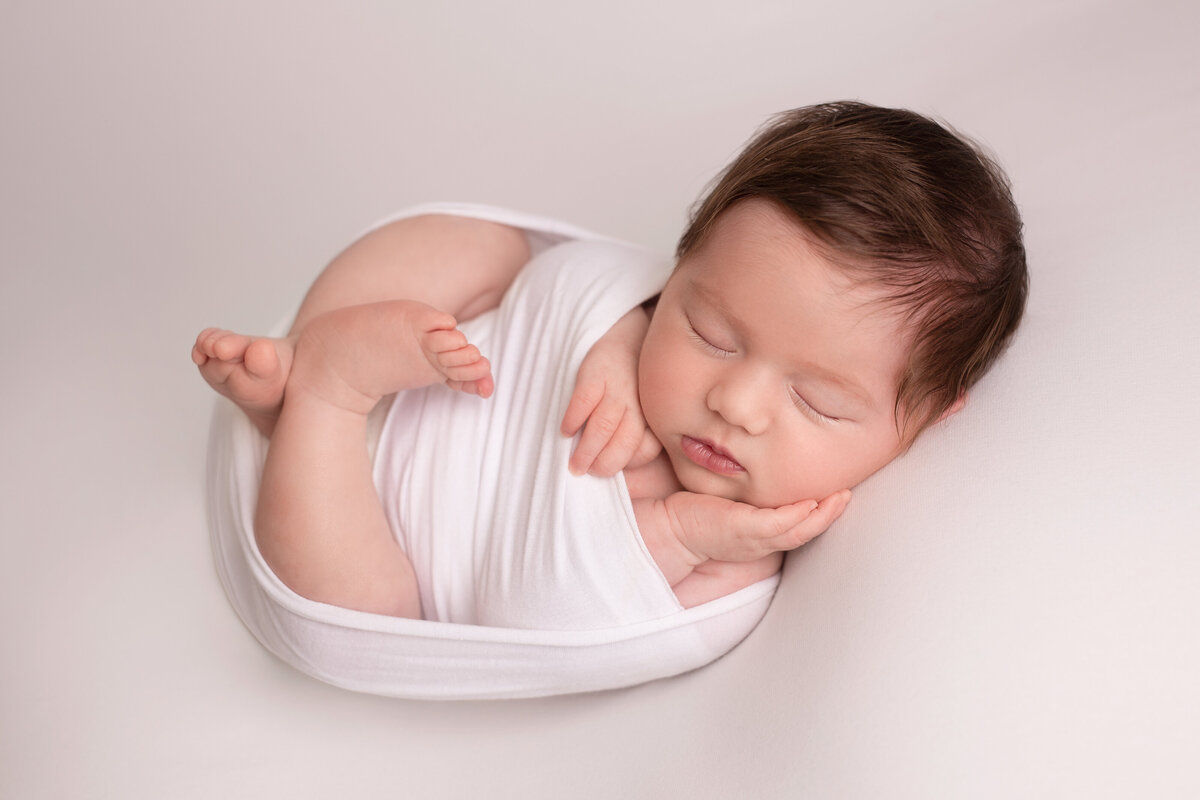 Newborn lying sleeping curled up wrapped on a white background with her hand on her cheek.