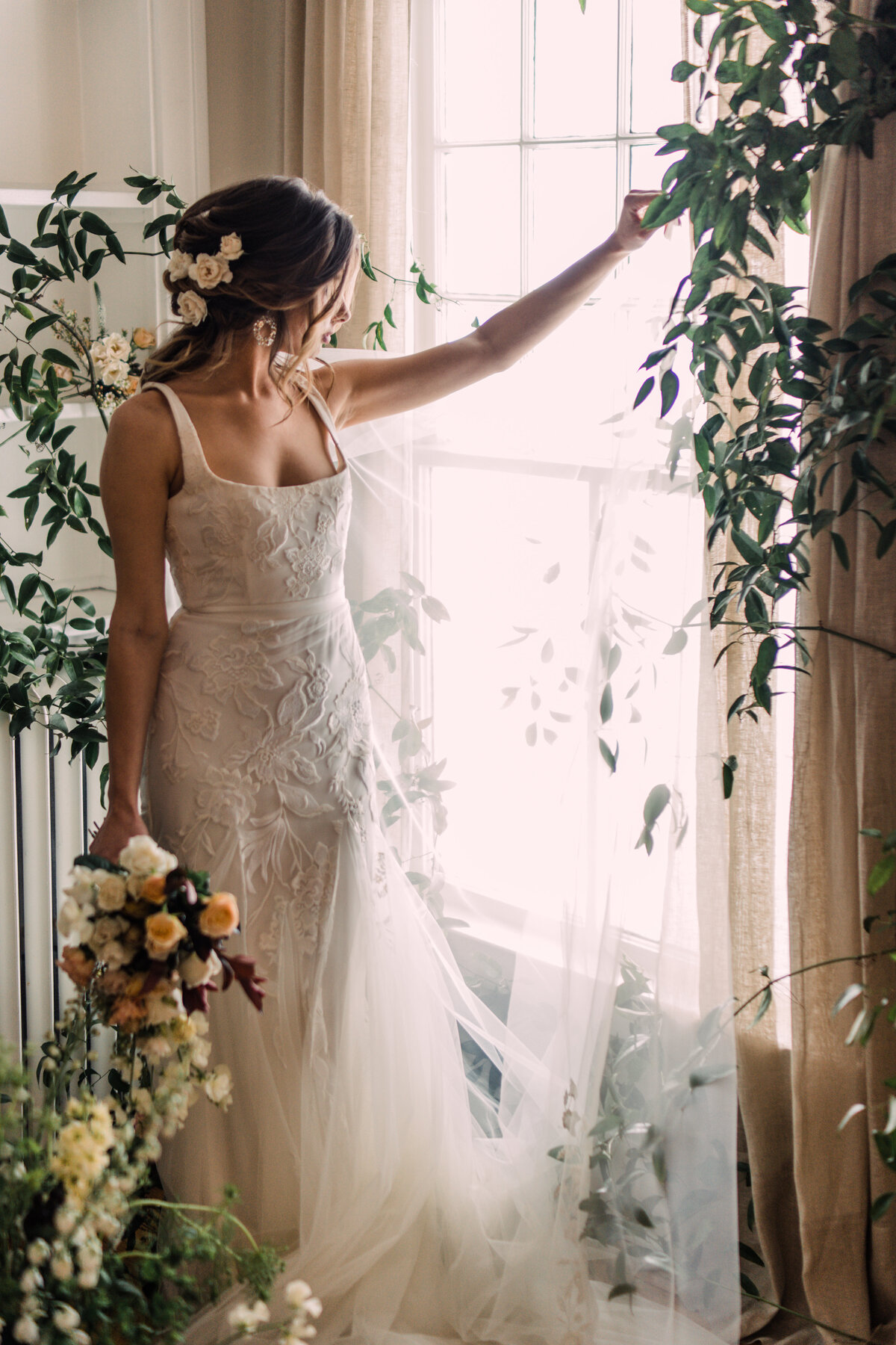 Bride wearing Alexandra Grecco Marcelle gown standing in front of window at Cornman Farms holding up Ainsley veil to the light