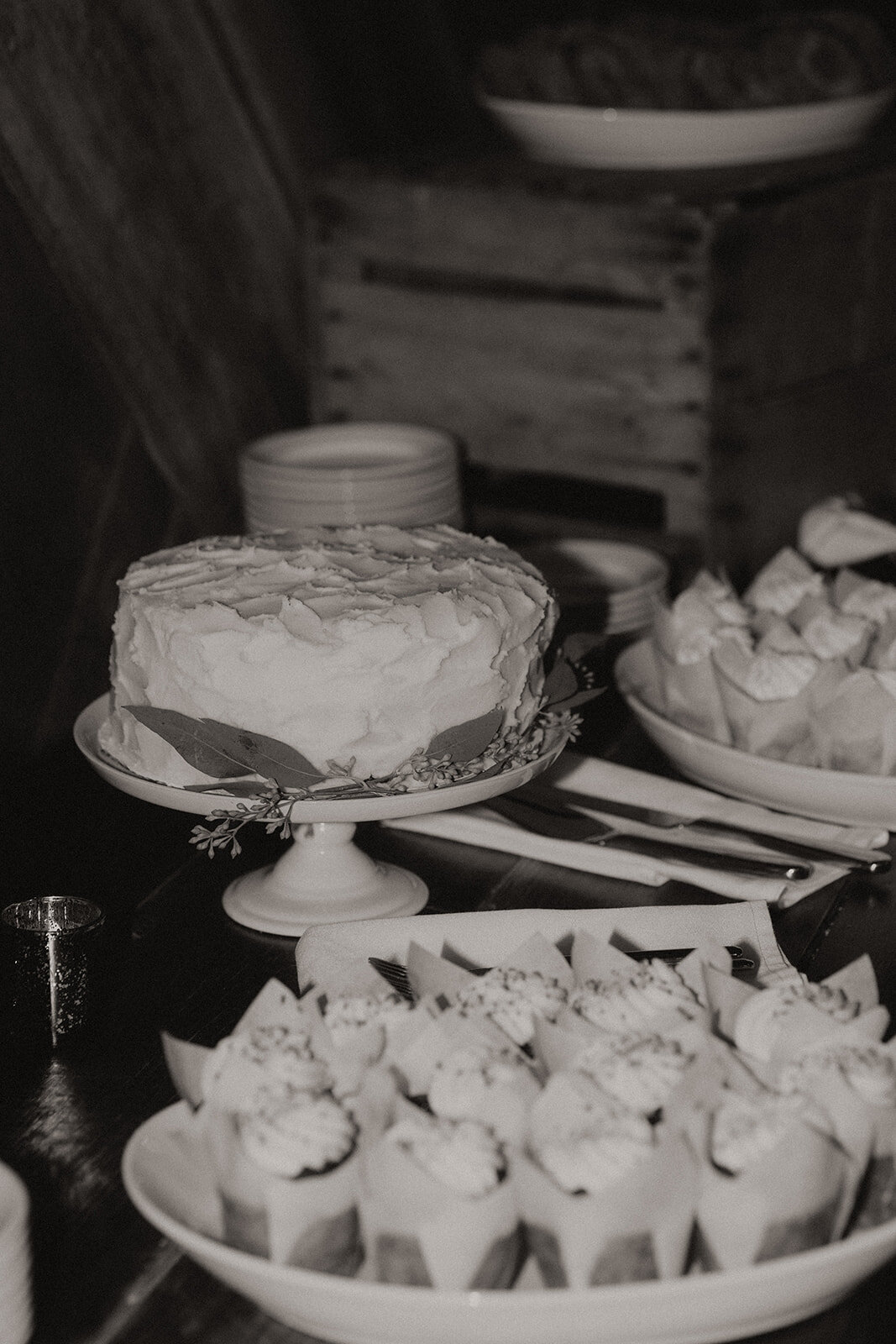 A black and white photo of a dessert table with a frosted cake and cupcakes, elegantly displayed on plates and stands with a rustic backdrop, organized by an Illinois wedding planner.
