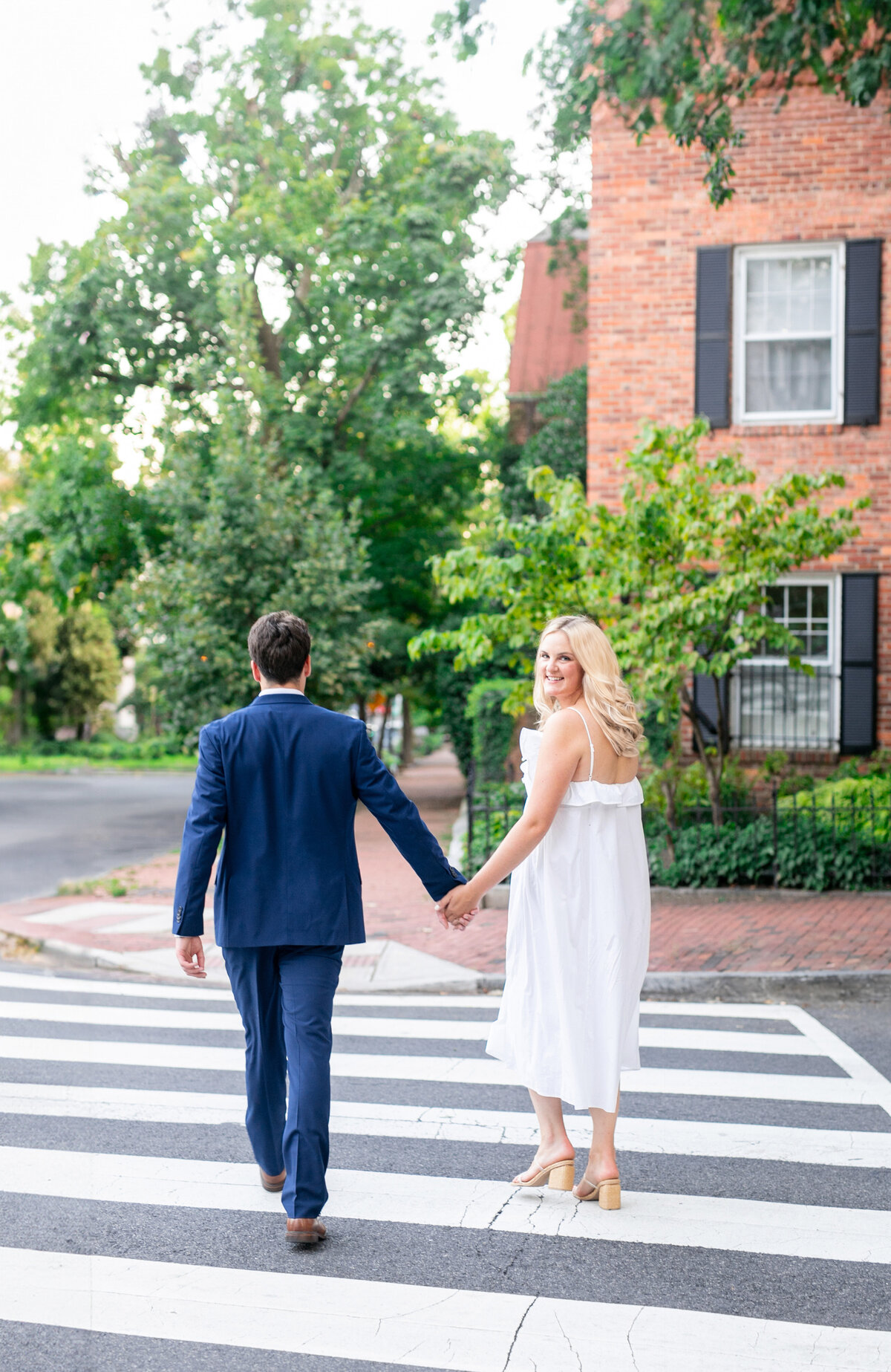 A couple holding hands walks across a zebra crossing. The man is in a blue suit, and the woman is in a white dress. The setting is a tree-lined street with a brick building in the background.