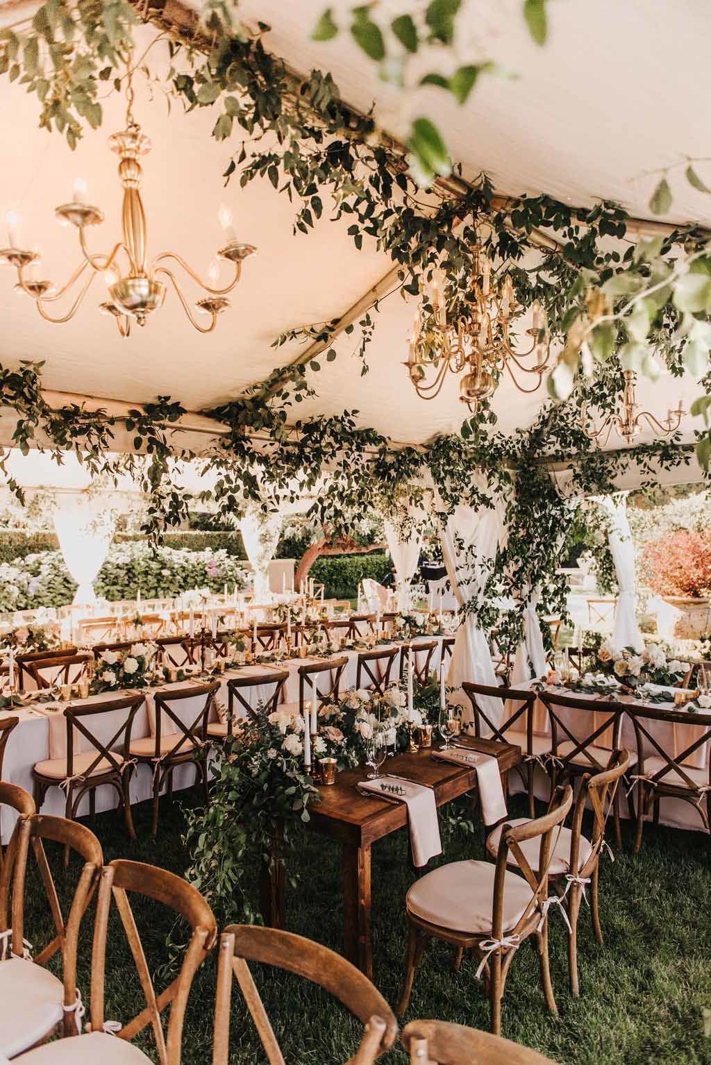 wedding reception tent covered in greenery with long wooden guest tables and vineyard chairs