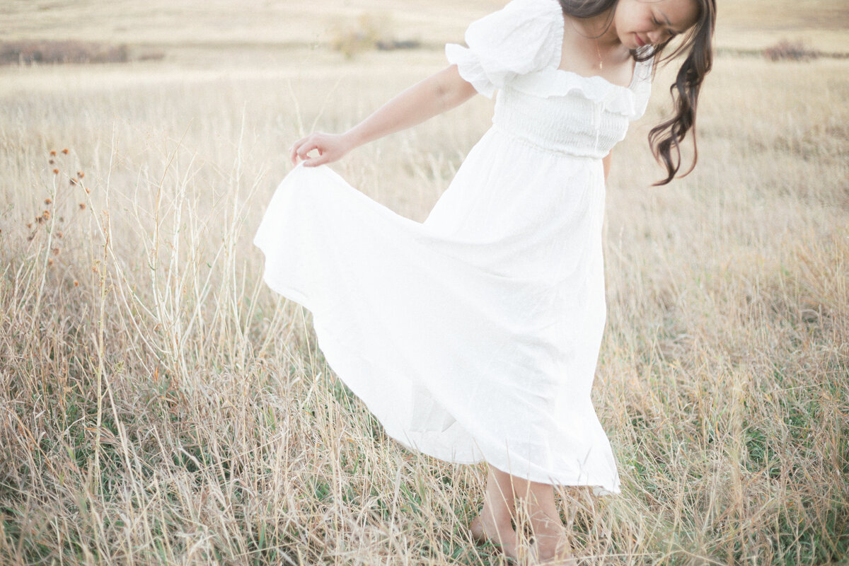 beautiful artistic photo of a mom in a long white dress dancing in a field. Mountains are in the background.
