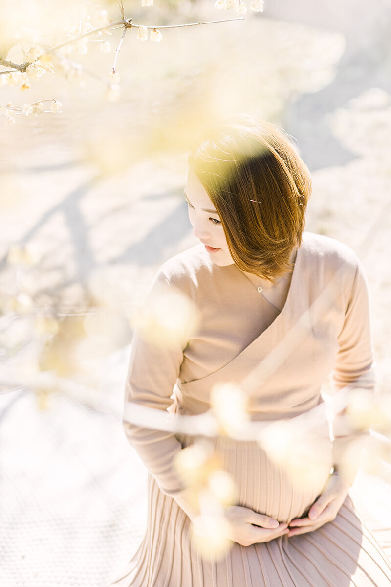 蠟梅の花の下でうつむきながら大きなおなかに手を添えた美しいマタニティフォト。町田市で撮影。