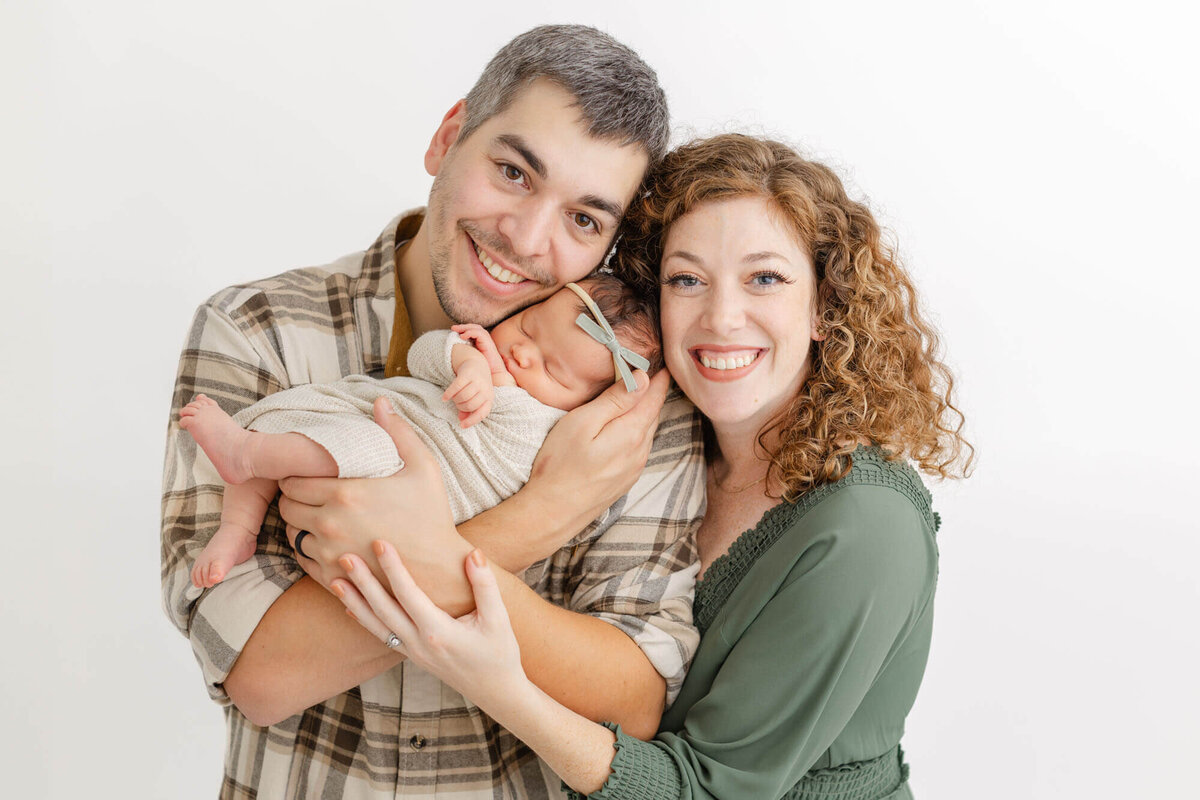 Dad holding newborn baby up to his cheek and mom is standing next to him at his shoulder snuggled up next to baby's head. Mom and Dad are both smiling at the camera