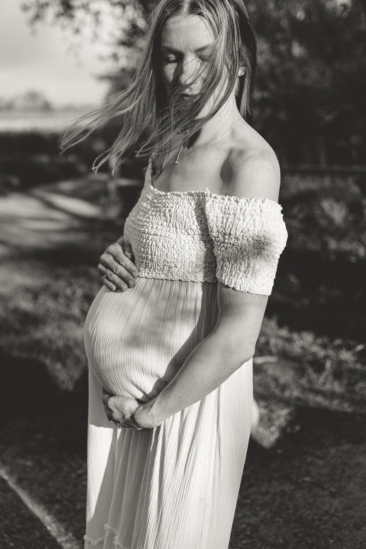 Mom holding pregnant belly wearing white dress with tree branch shadows casting on gown and hair flowing in the wind during session with alexis adkins photography in firestone colorado