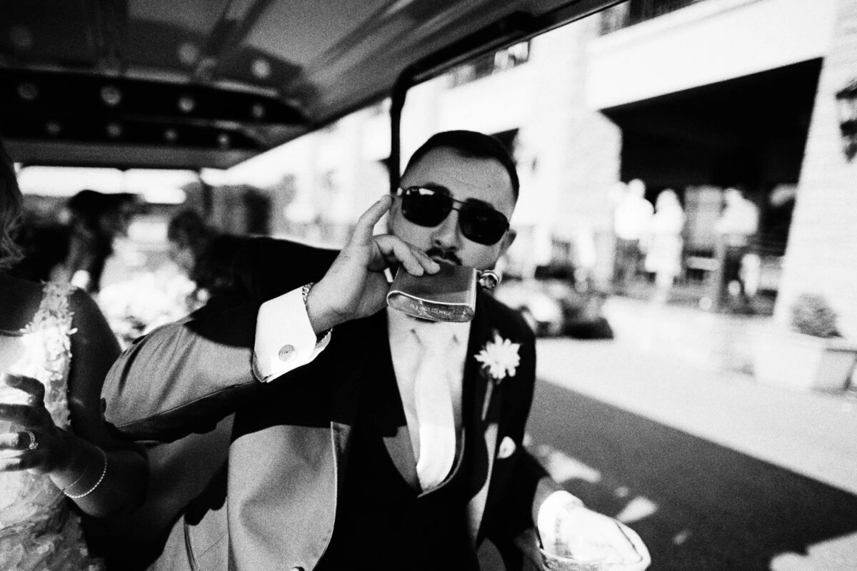 A man wearing a suit and sunglasses holds a drink and gestures toward the camera while riding in a vehicle. Captured by a film photographer NJ, the black-and-white image highlights the flower pinned to his lapel.
