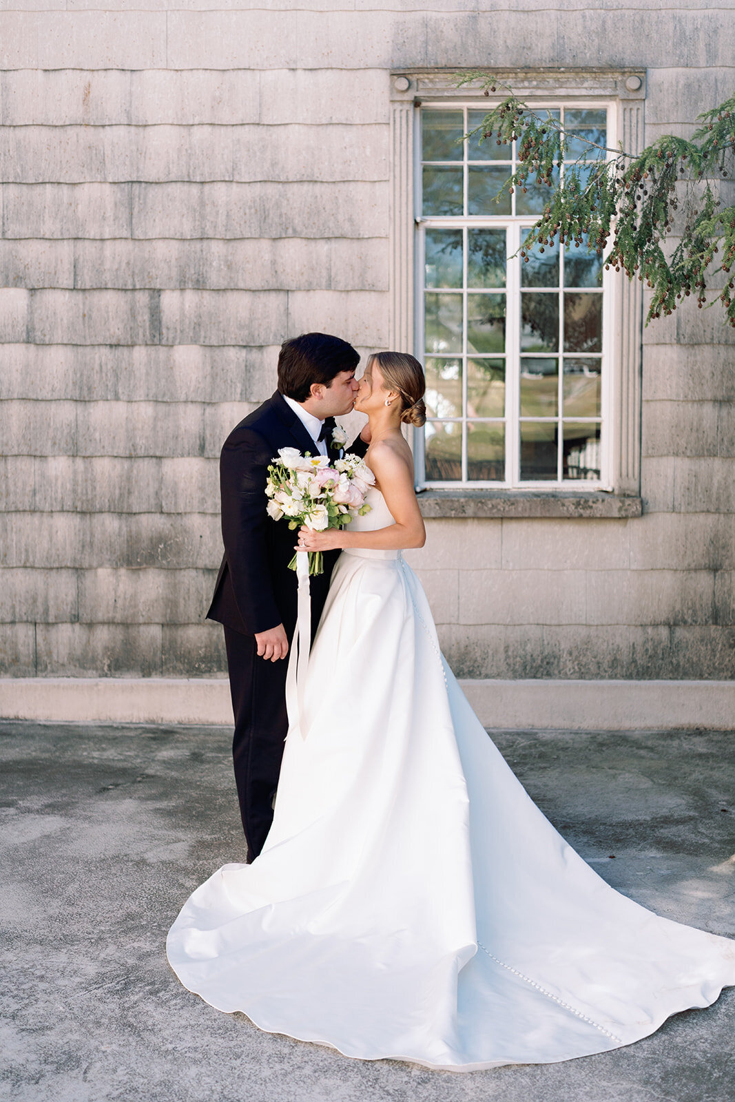 Bride and groom sharing a kiss while holding a bouquet in front of the stone wall at Burritt on the Mountain in Huntsville, Alabama, photographed by Perry Hancock.