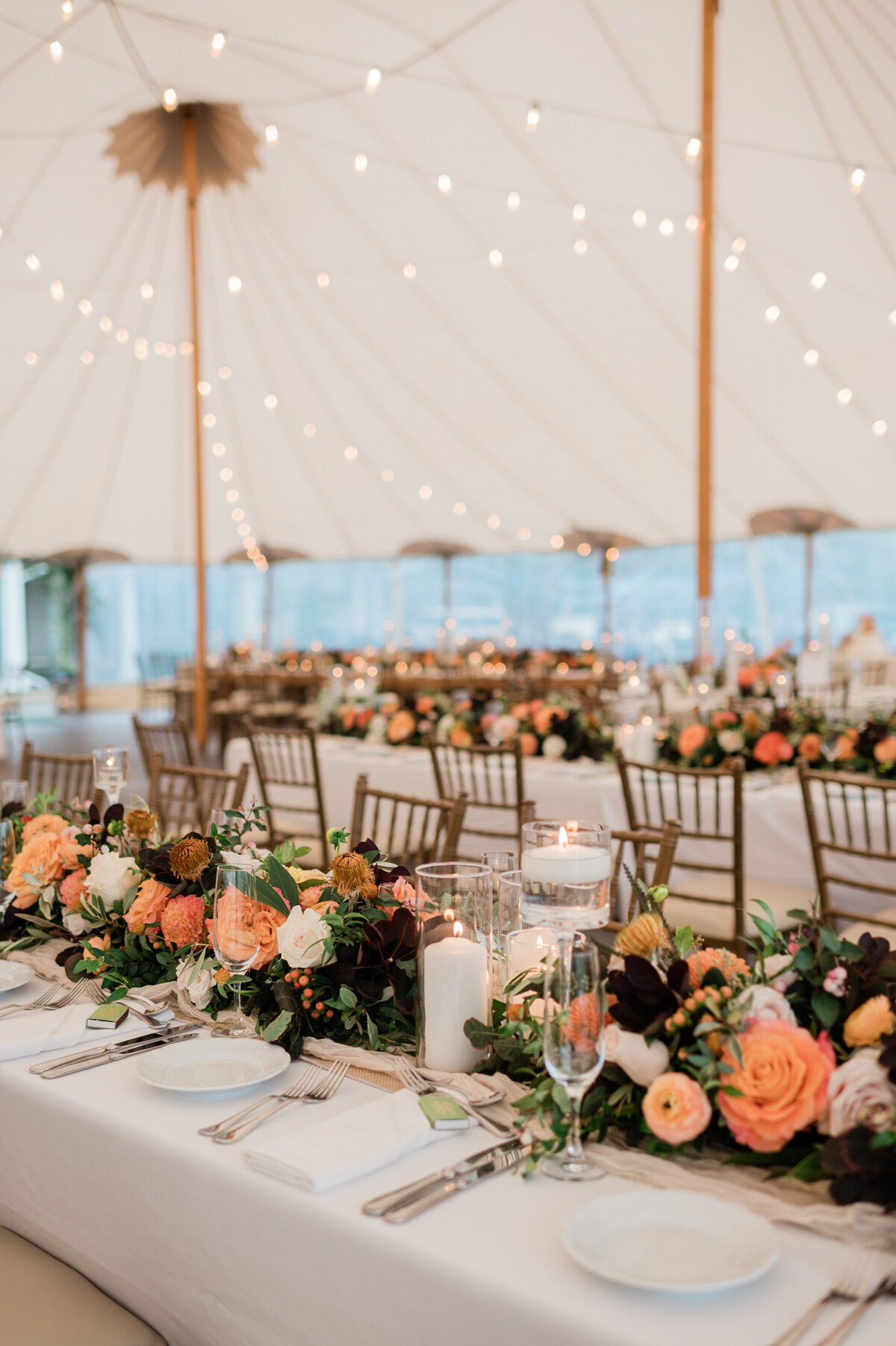 A long, white-draped dining table is decorated with a floral garland featuring peach and cream roses, greenery, and floating candles.