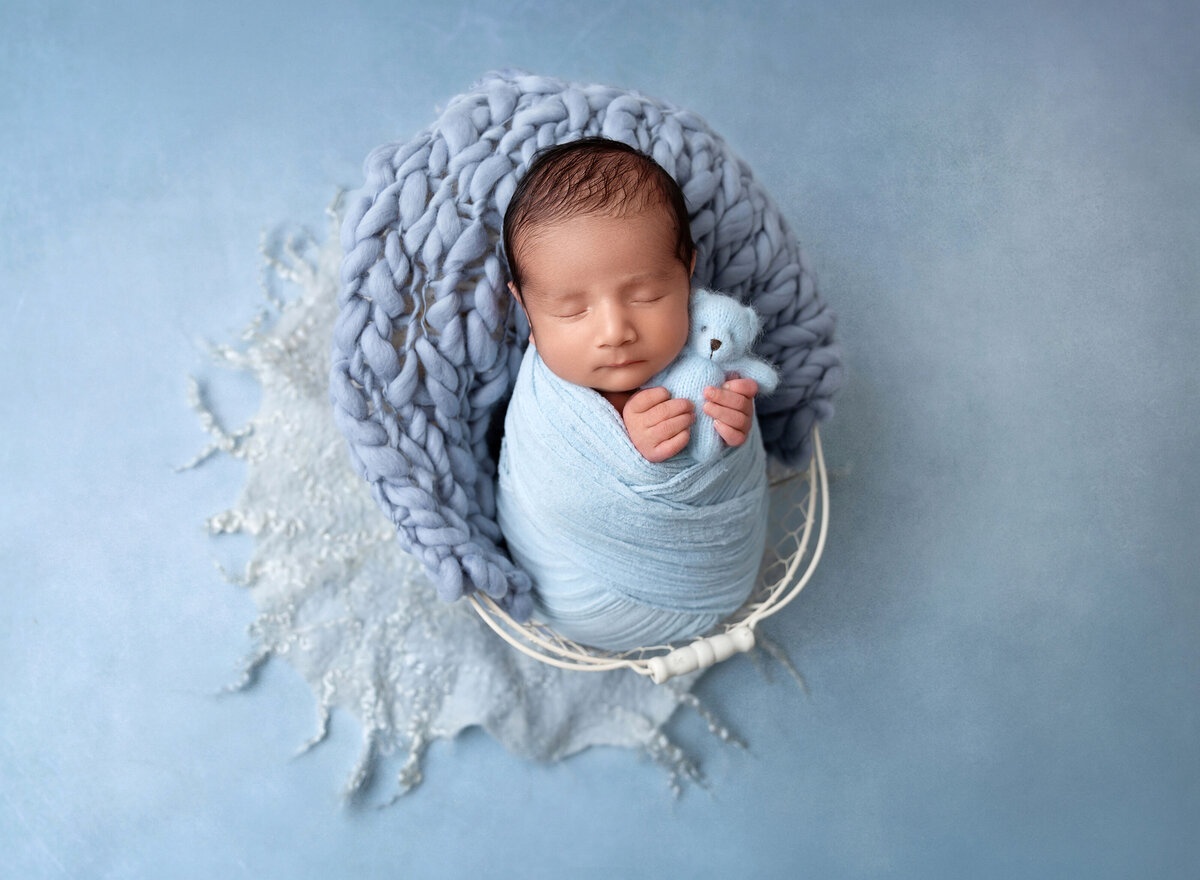 Baby boy is swaddled with his hands peeking out holding a petite light blue knit teddy bear. Baby is sleeping.