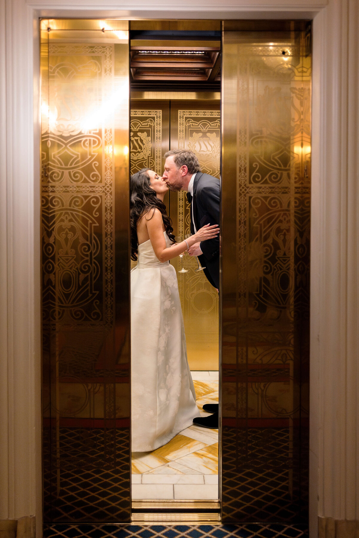 A couple shares a tender kiss inside a lavish elevator with ornate golden designs on the doors. The woman is in a white dress, and the man is in a dark suit. The scene is filled with an atmosphere of elegance and romance.