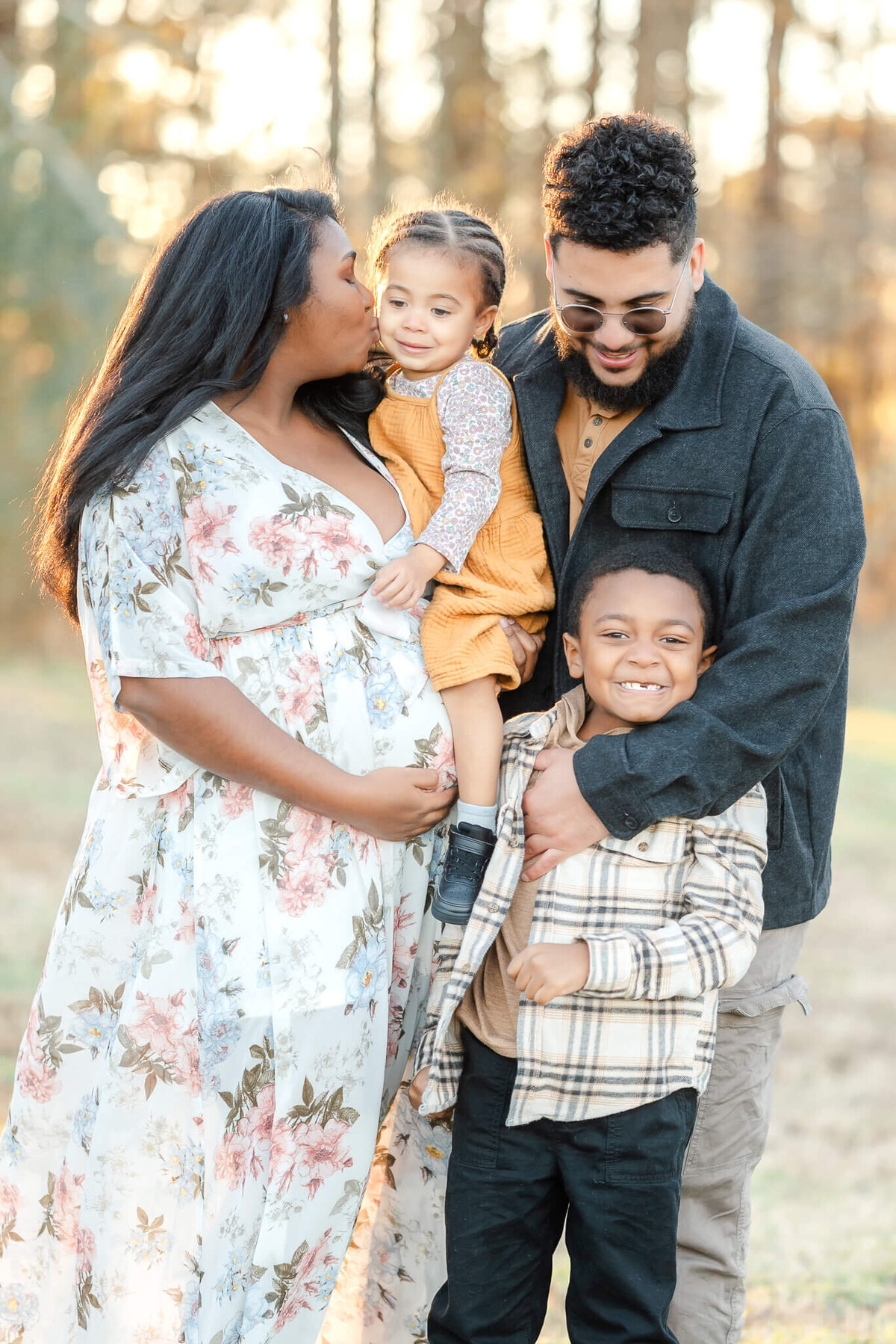 A family snuggles close during a maternity session in Chesapeake, VA. Everyone has their arms around each other and the pregnant mama kisses her toddler.