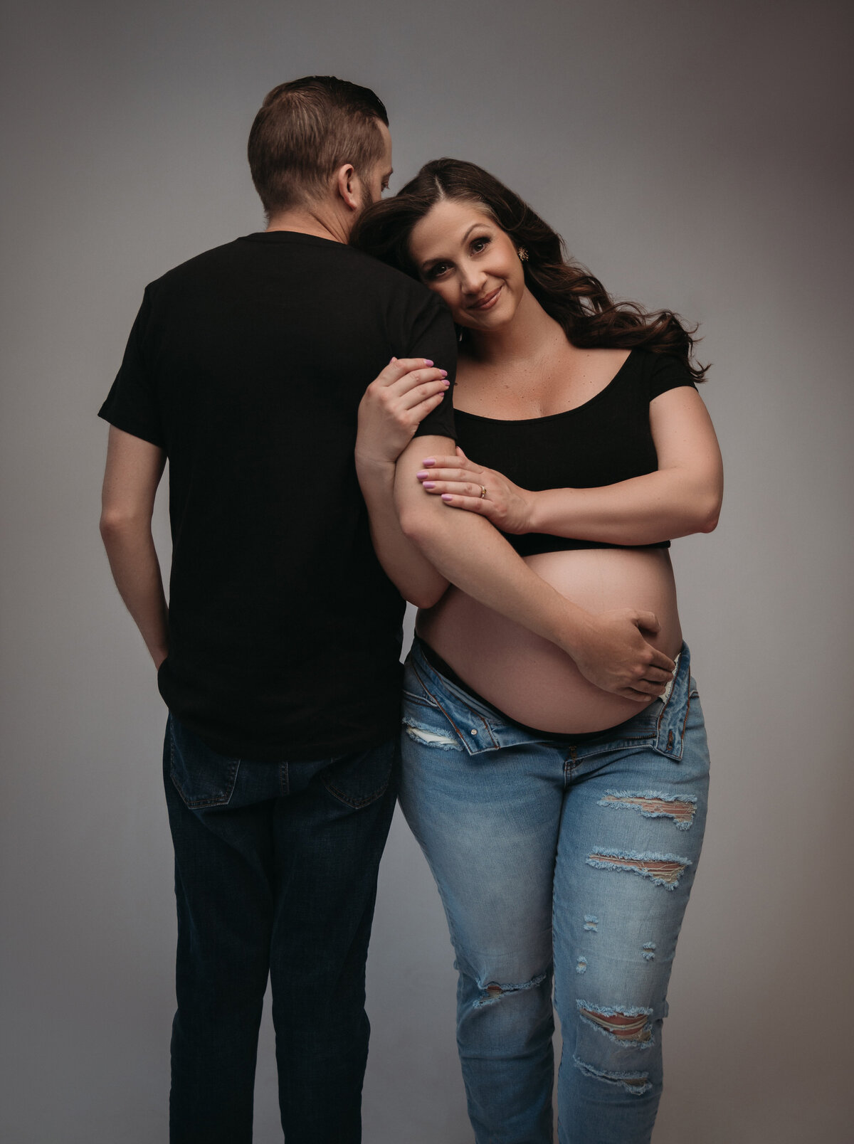 dad to be wearing dark jeans and shirt looking back while holding pregnant wife's baby bump while she holds his arms and looks at the camera and also wearing jeans and  a radk crop top