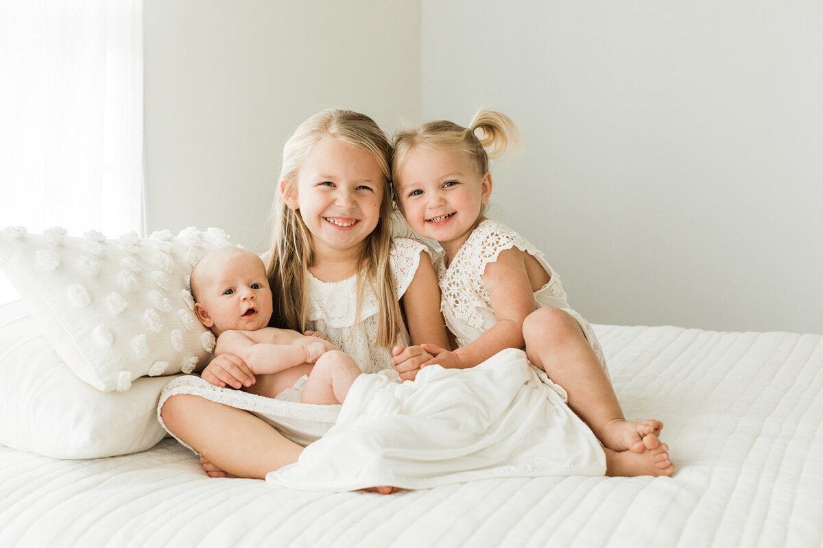 two blonde big sisters wearing white sit on a white bed holding their new little sister smiling