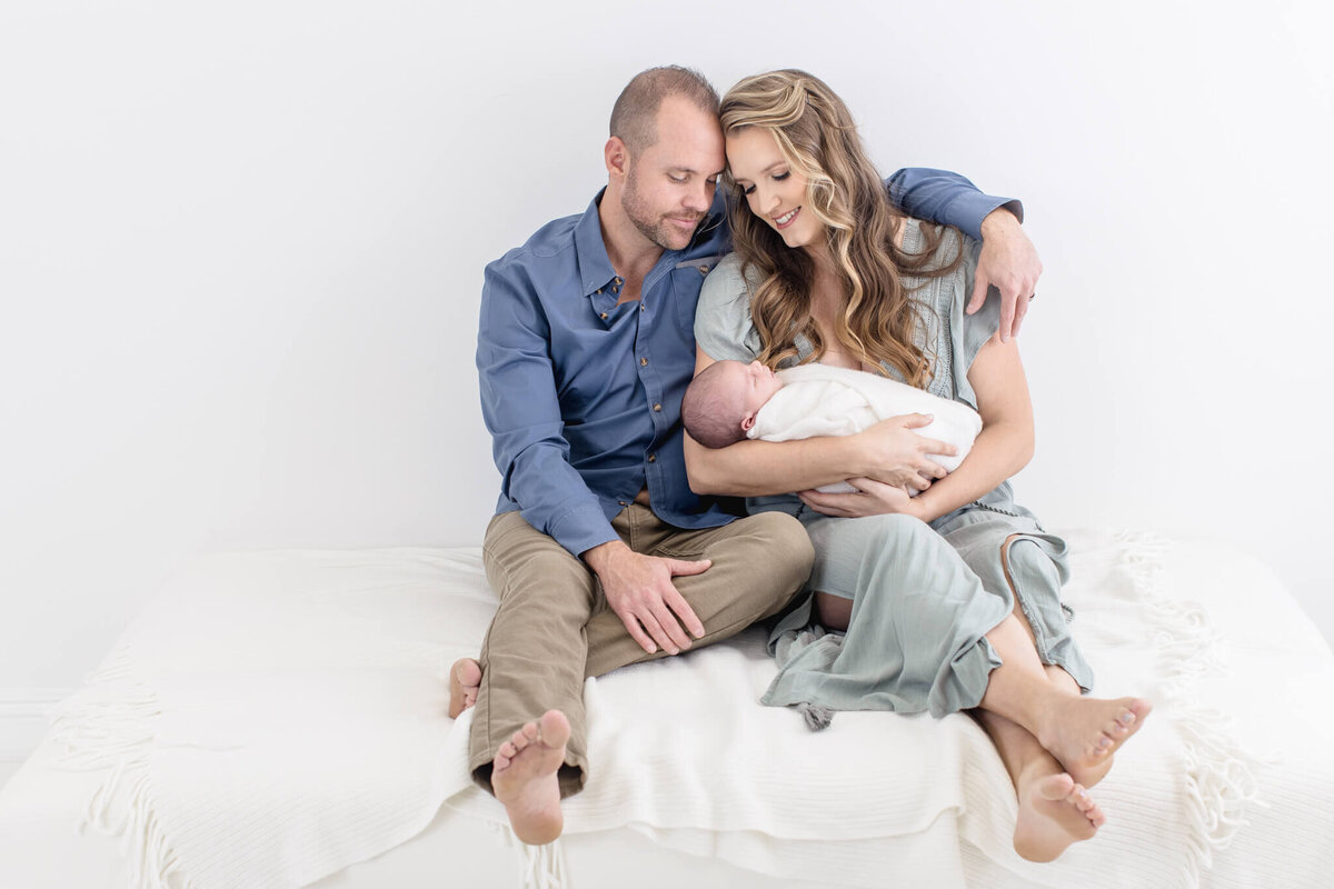 A couple sits closely on a soft, white bed, the man in a blue shirt and khaki pants, the woman in a light dress. She gently cradles a newborn baby wrapped in a white blanket, both parents looking lovingly at the infant.
