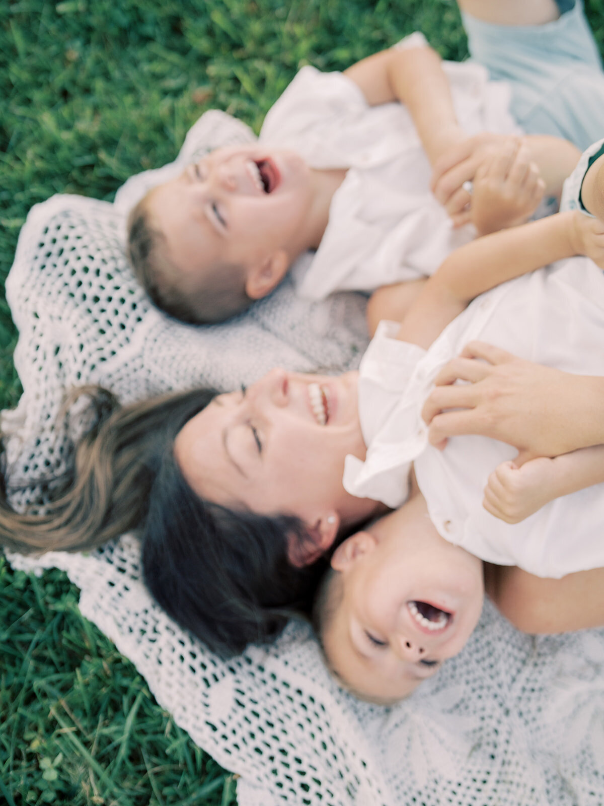 Mother with brown hair lays on crotchet blanket laughing with her twin boys.