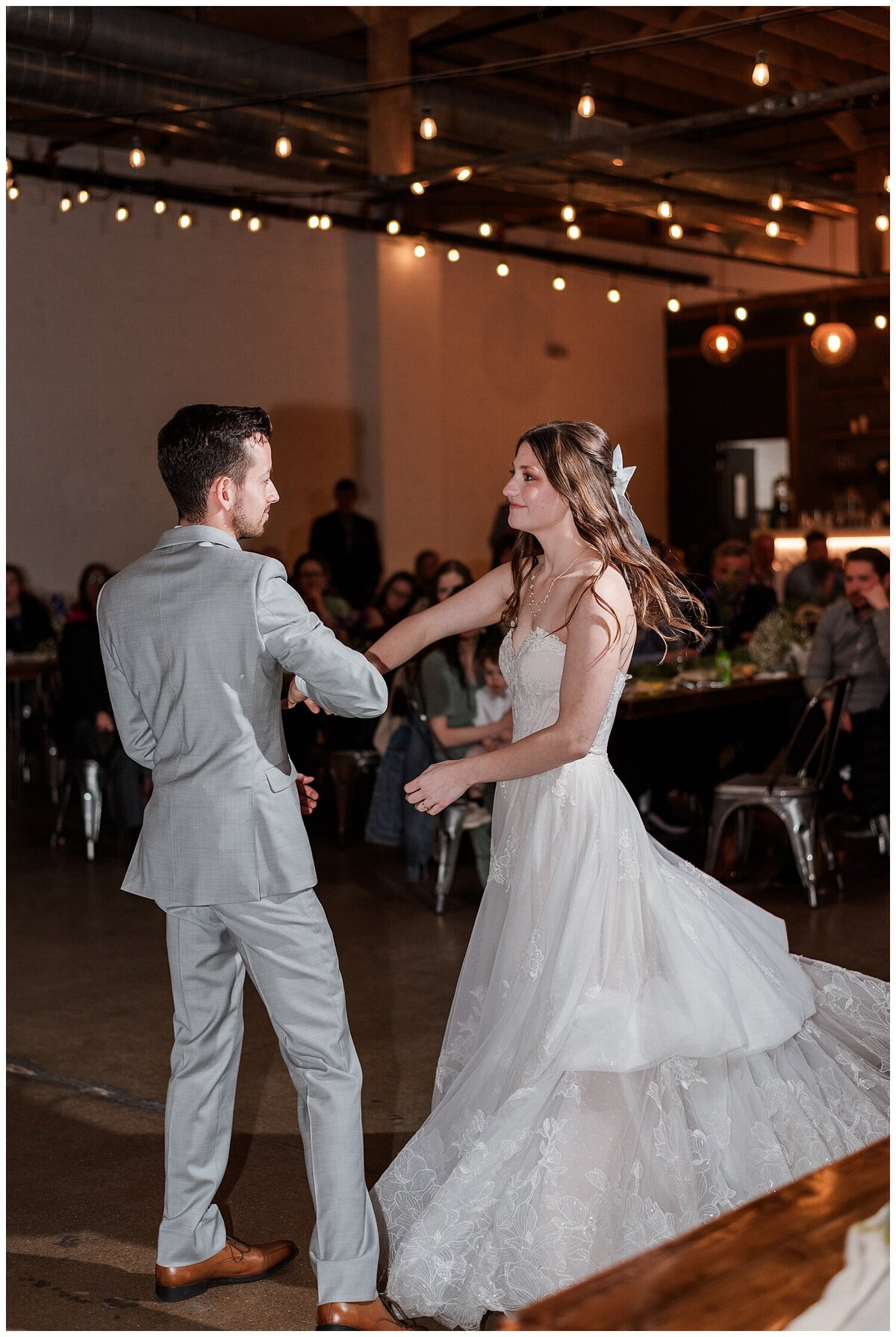 A groom in a light gray suit dances with his new wife and spins her as her dress flares out all around her and they look right at each other while at their venue skylight in downtown denver